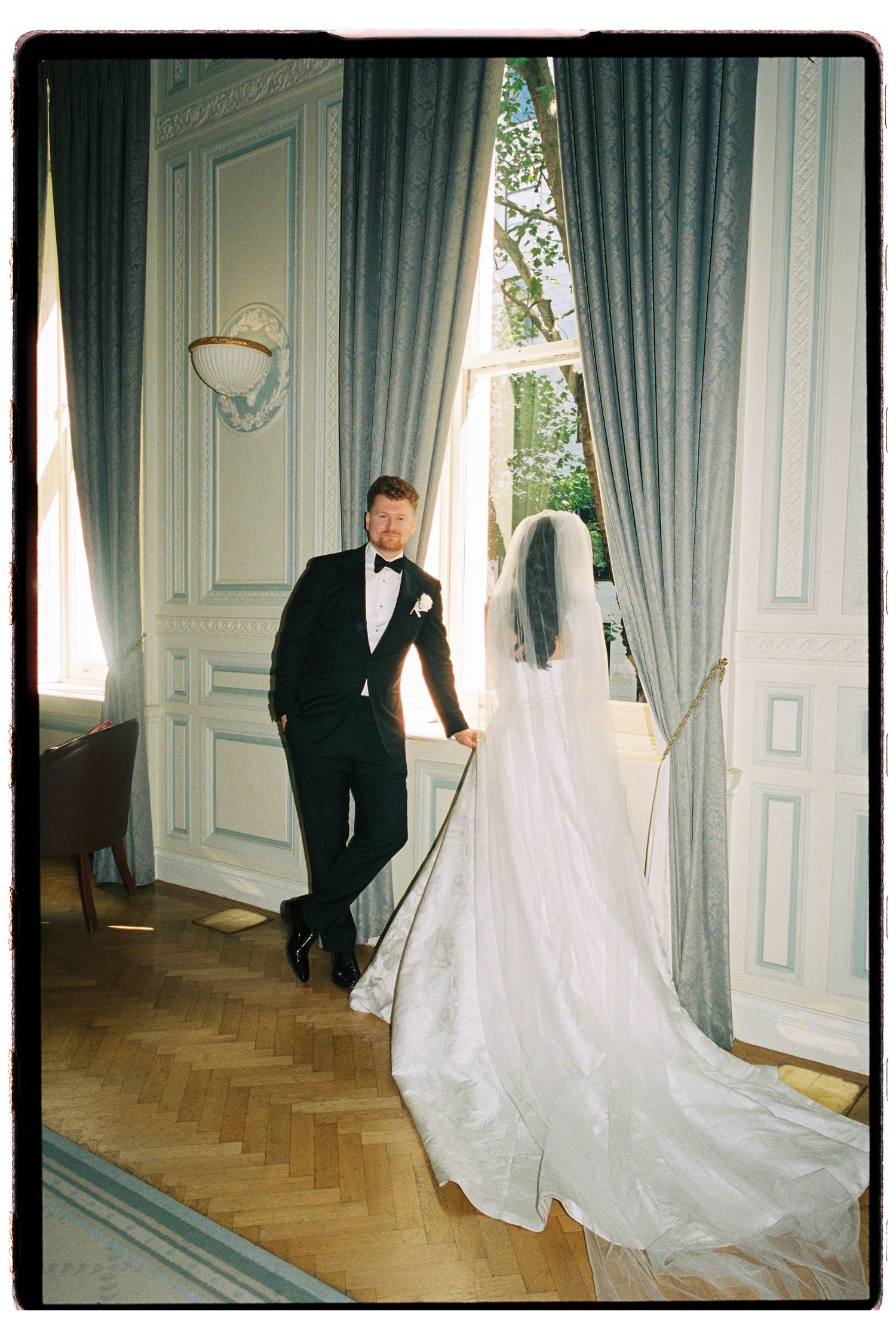 Film photo of groom leaning against window as bride in flowing veil stands in ornate French room.