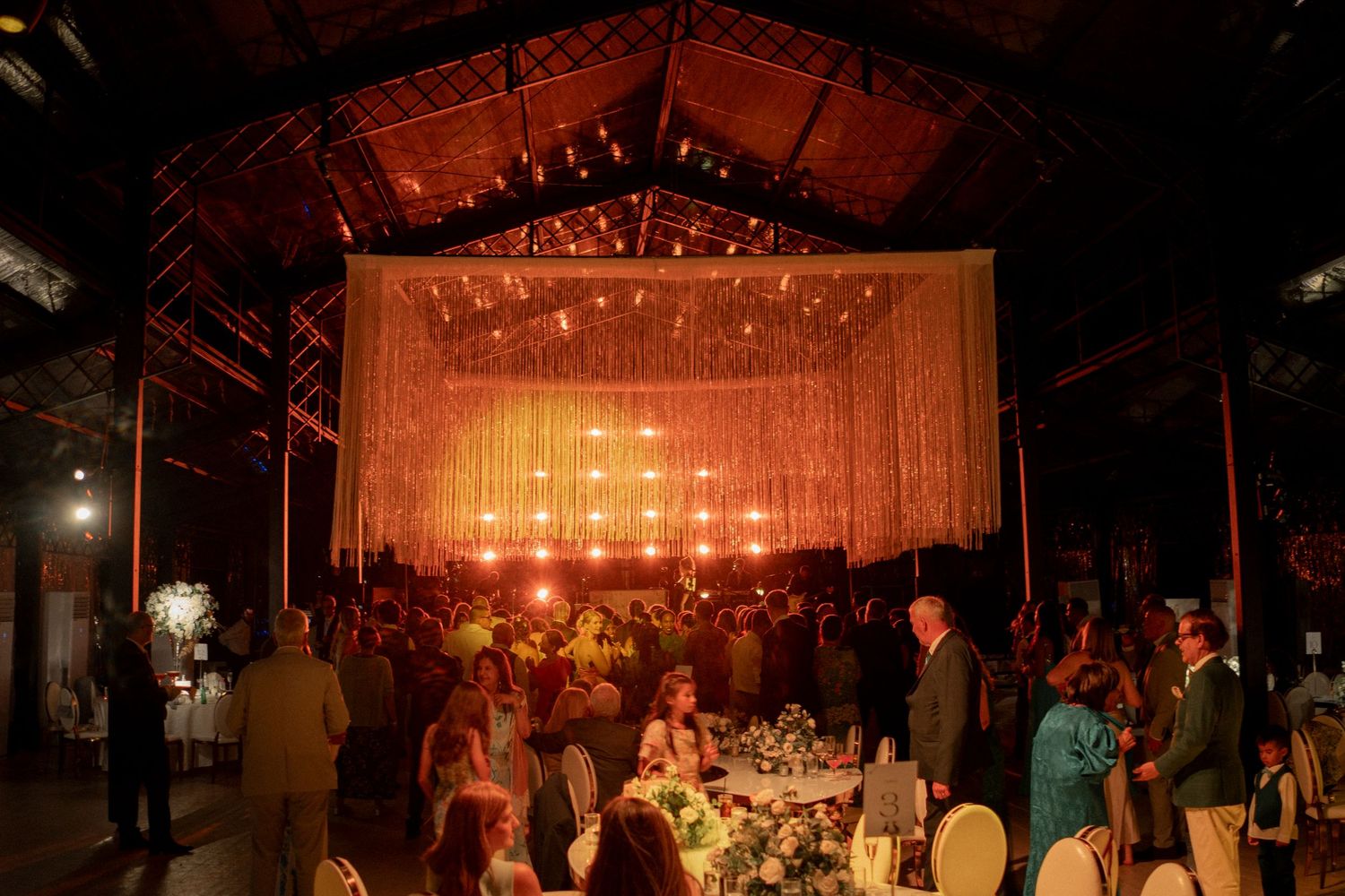 Guests dining at a romantic wedding reception in a warmly lit barn venue with string lights.