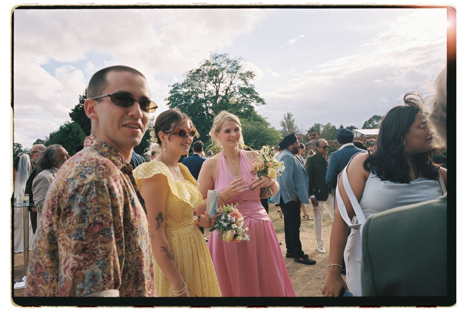 Guests in yellow and pink dresses holding bouquets at an outdoor wedding celebration on a cloudy day.