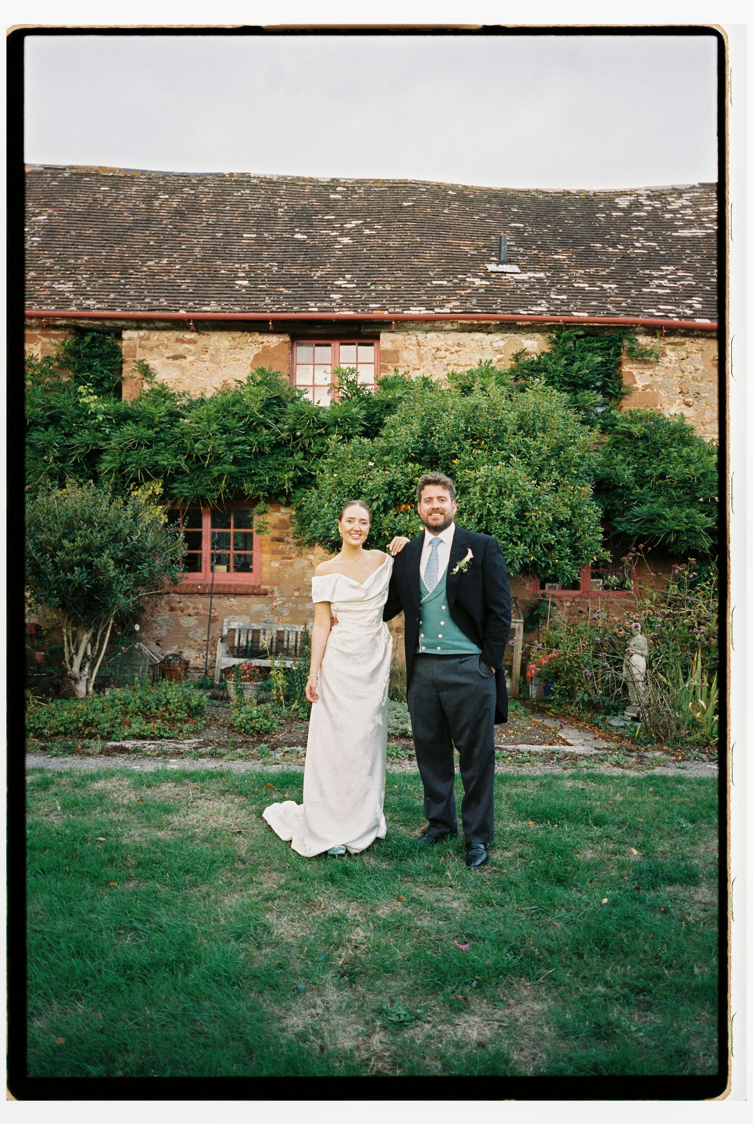 Bride and groom posing together on lawn in front of a rustic stone farmhouse wedding venue.