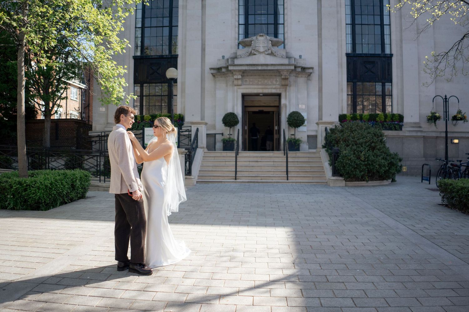 Bride and groom dancing in front of an elegant building on their wedding day in a sunny outdoor plaza.