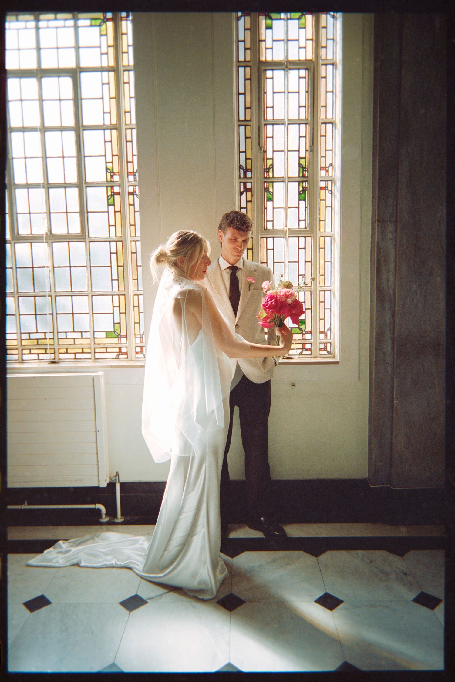 Bride and groom share a tender moment by stained glass windows, bride in flowing white gown with veil holding pink bouquet.