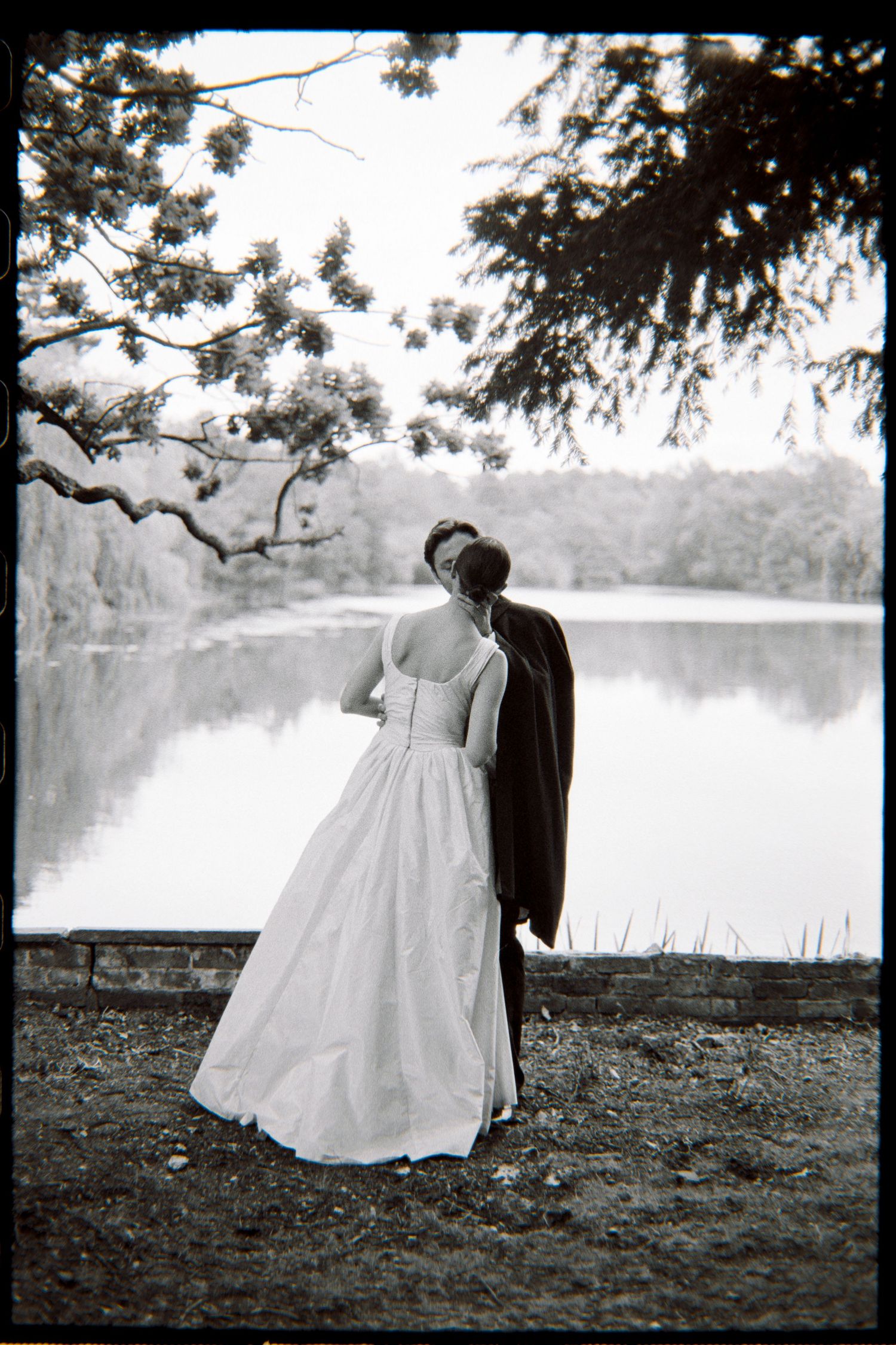 Black and white photo of a bride and groom sharing a kiss by a serene lake, framed by overhanging tree branches.