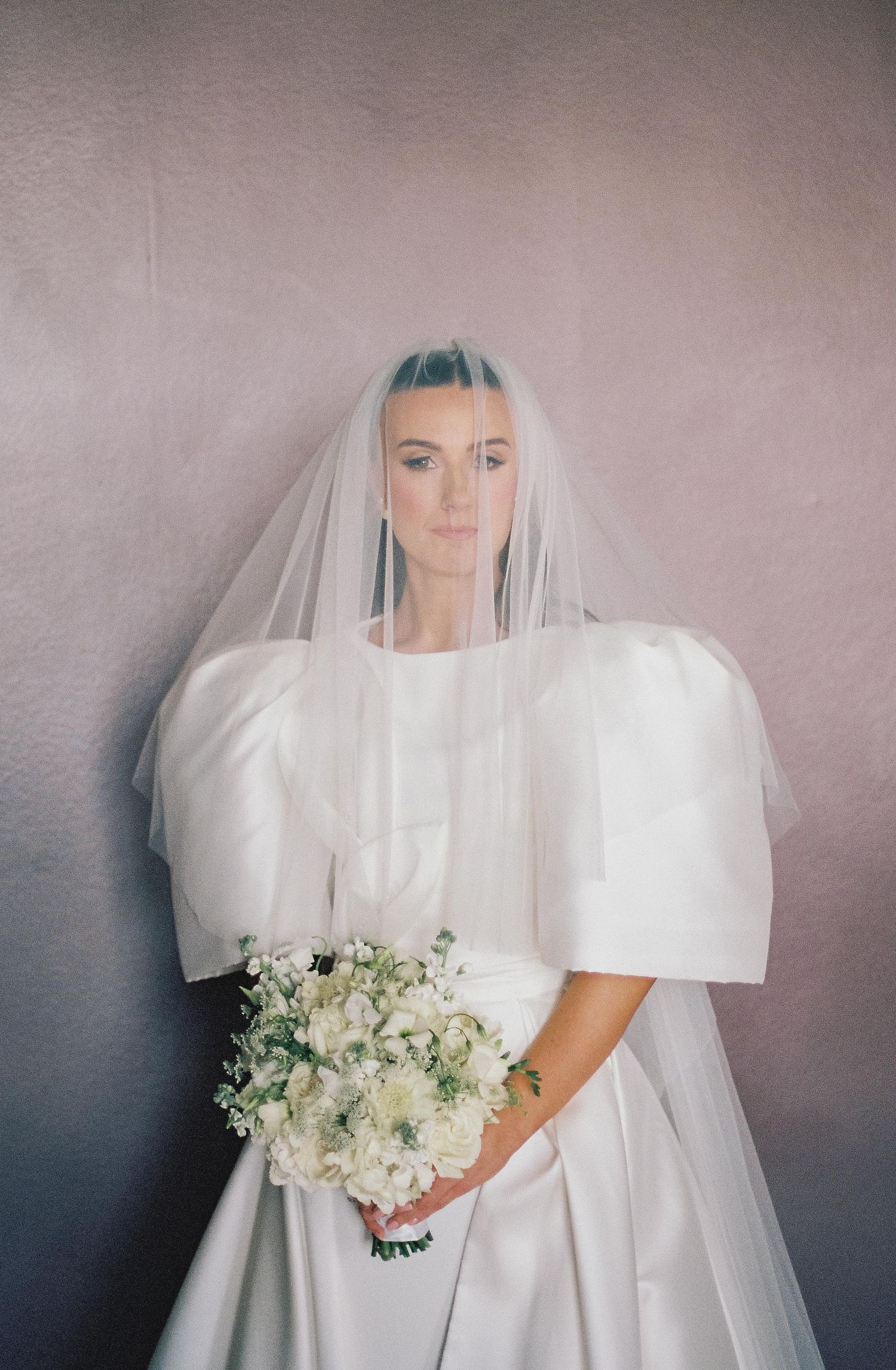 Modern bride in structured white wedding gown with puff sleeves and veil holding classic white bouquet.
