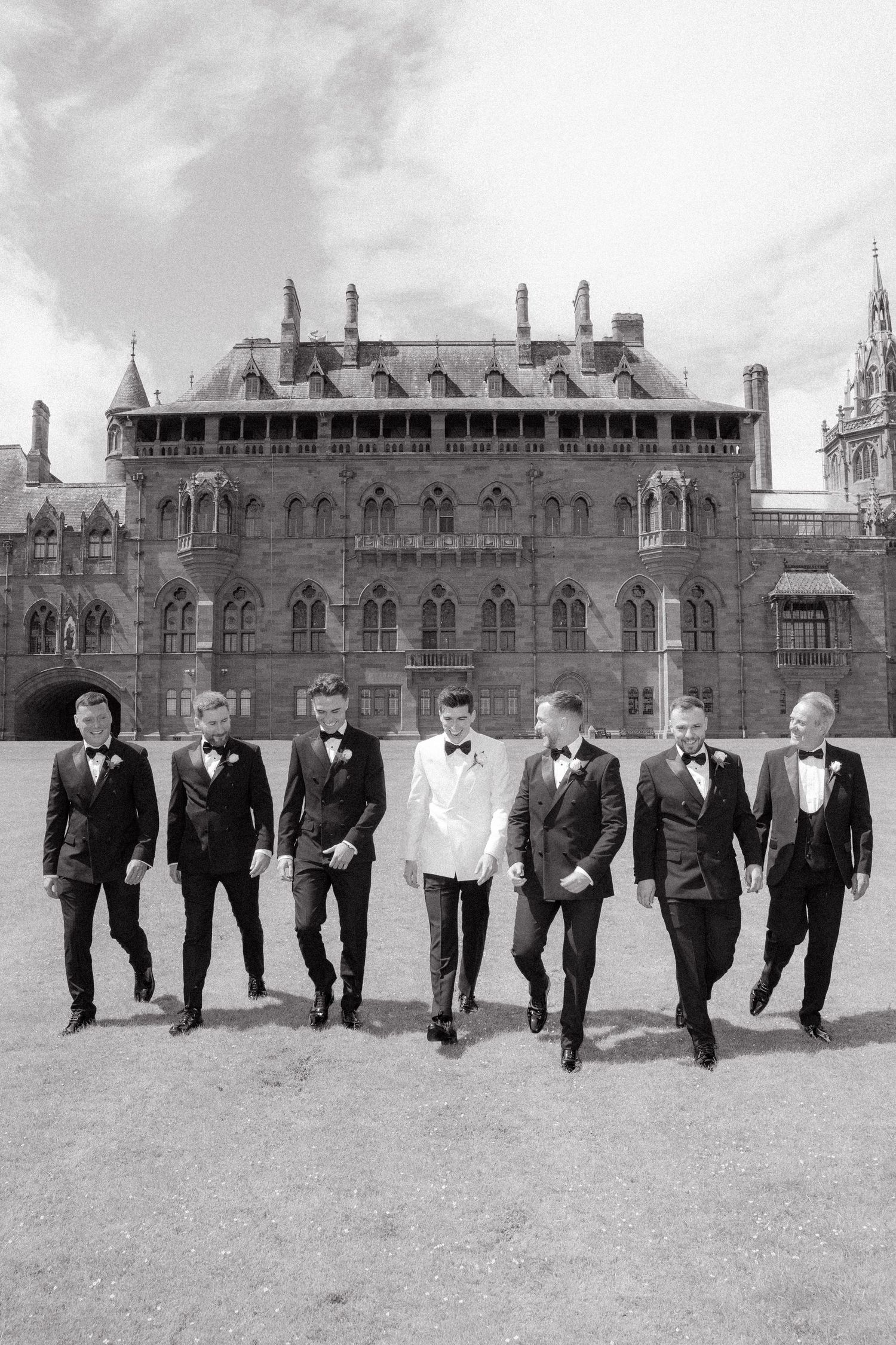 Groomsmen in black tuxedos walking in formation against a historic stone building backdrop.