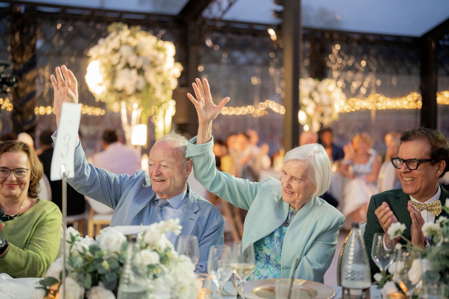 Elderly couple joyfully raises hands at wedding reception dinner with white floral centerpieces and string lights.