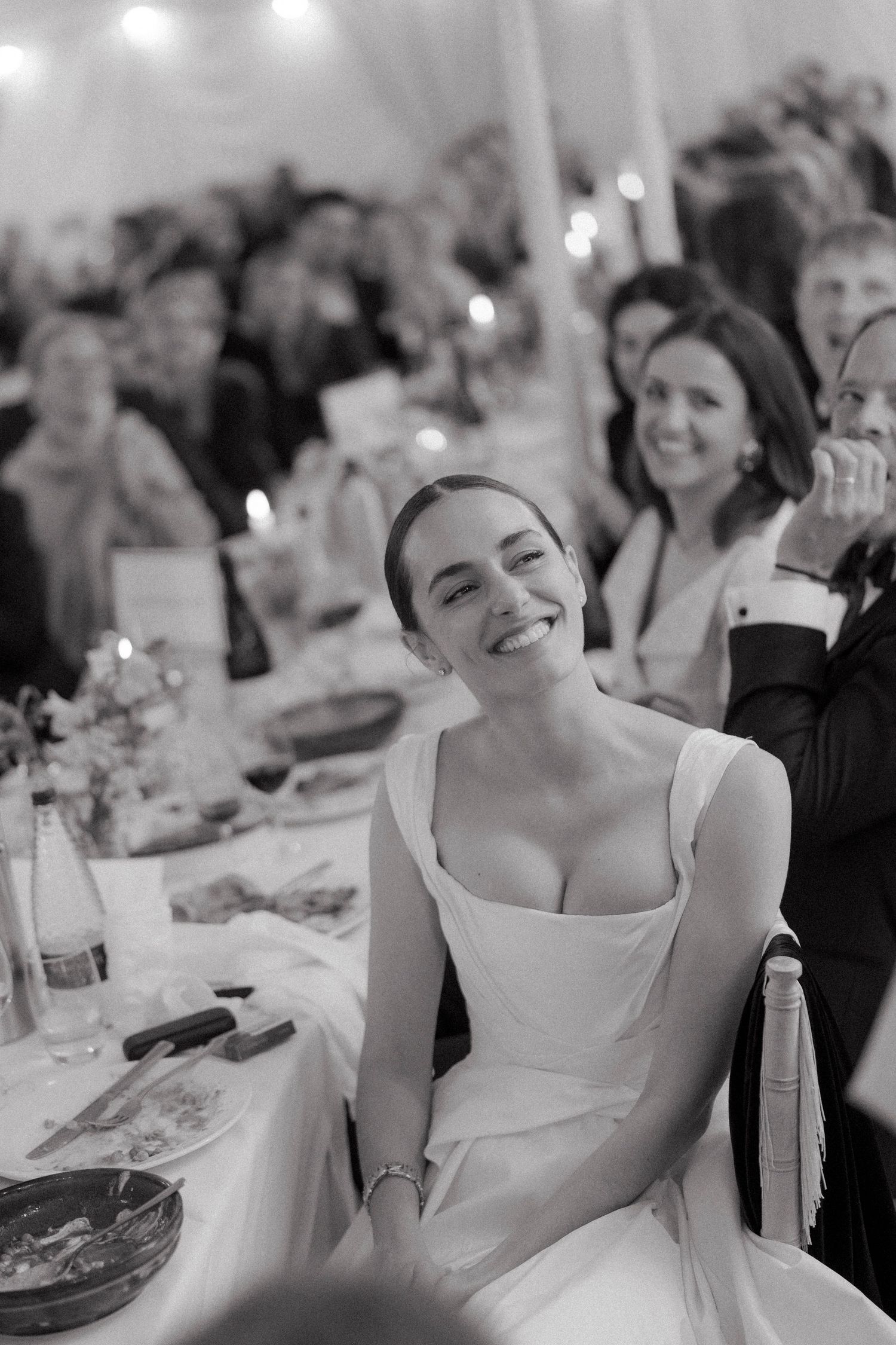 Smiling bride in a white gown with a square neckline seated at a candlelit reception table, with joyful wedding guests behind her.