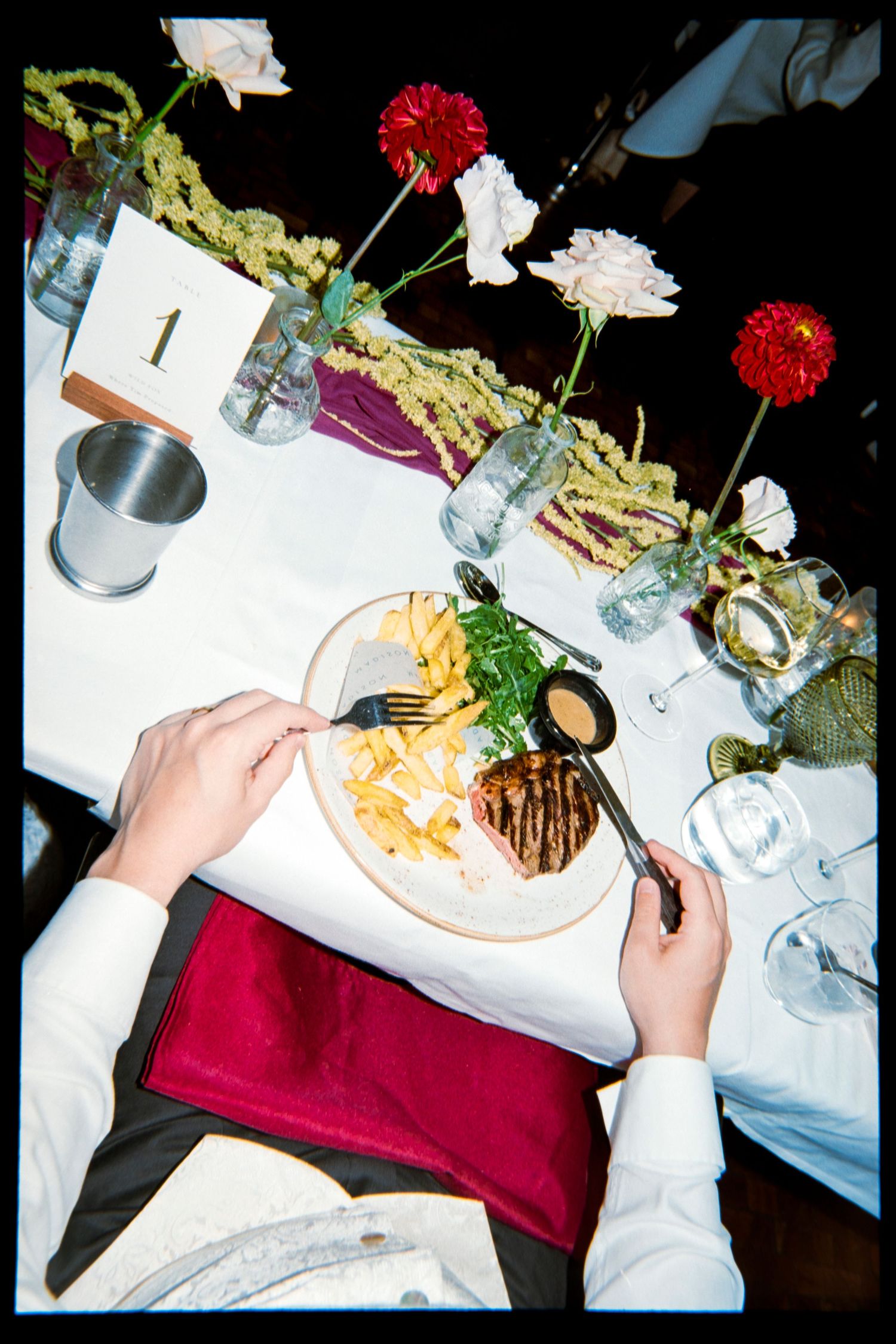 Overhead view of a diner cutting steak with fries and greens at table number 1, set with red dahlias, white roses and a burgundy napkin.