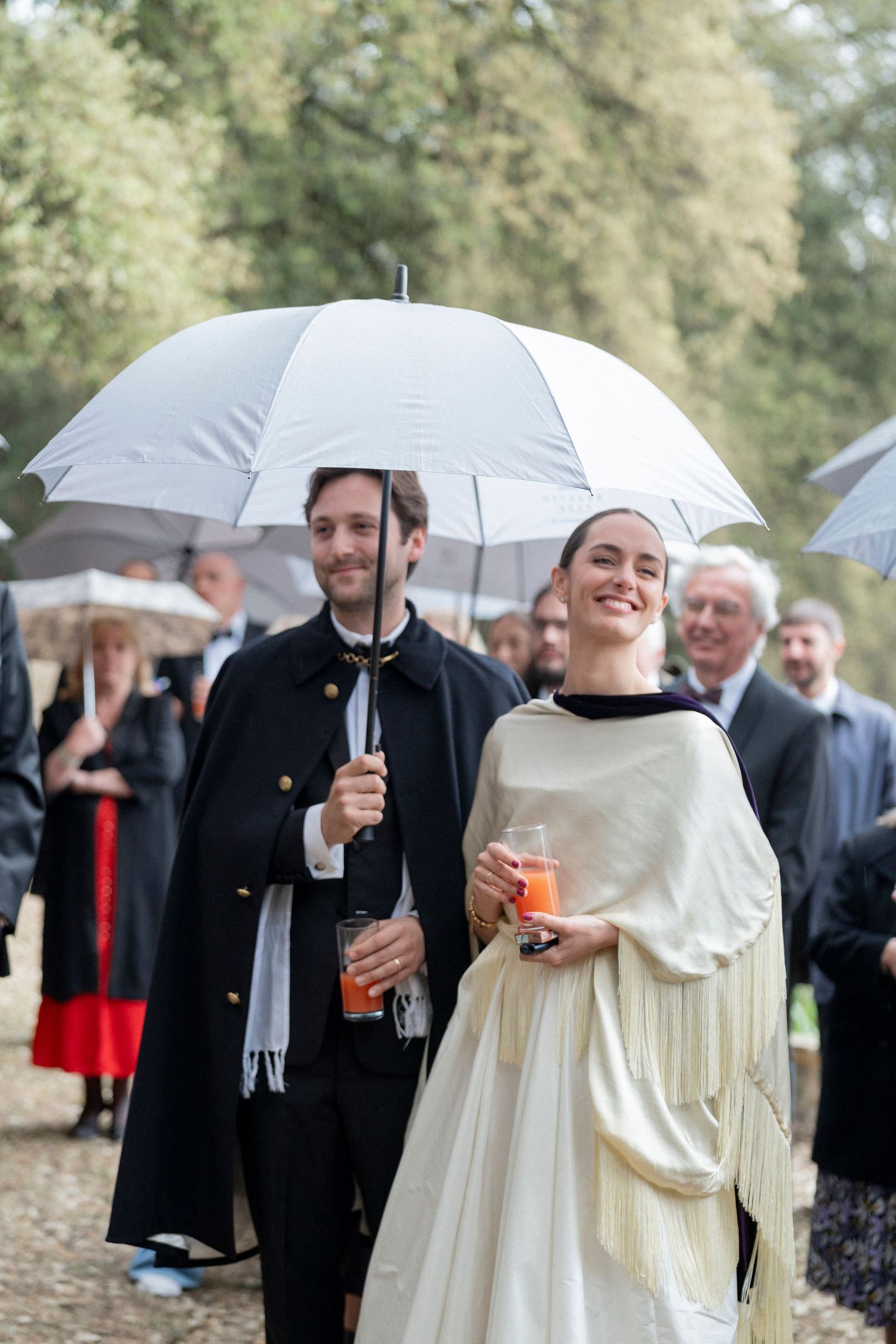 Smiling couple in formal attire hold orange cocktails under a white umbrella as wedding guests gather outdoors in the rain.
