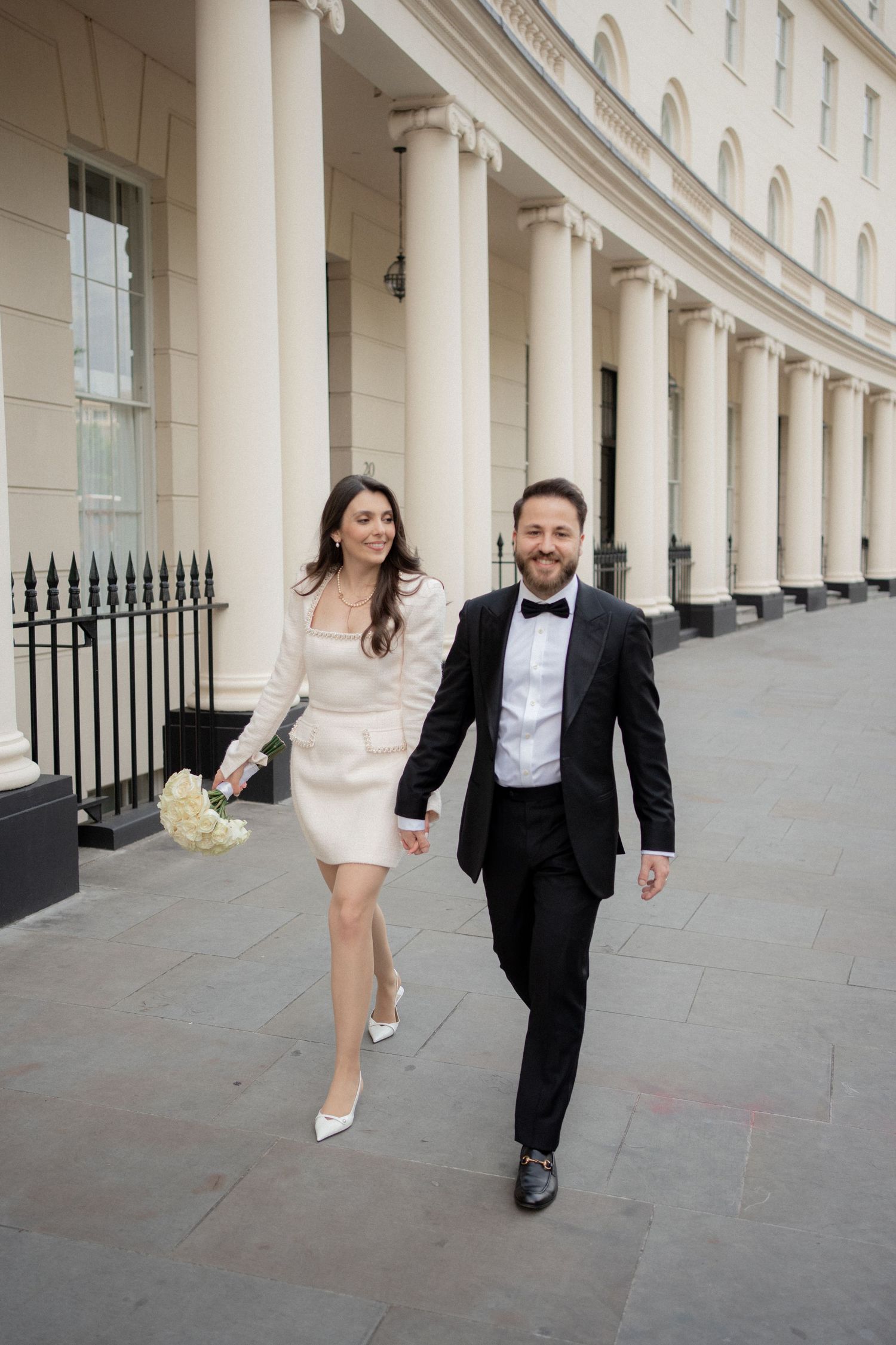 A couple in formal attire runs playfully along a grand colonnade with white pillars and iron railings.