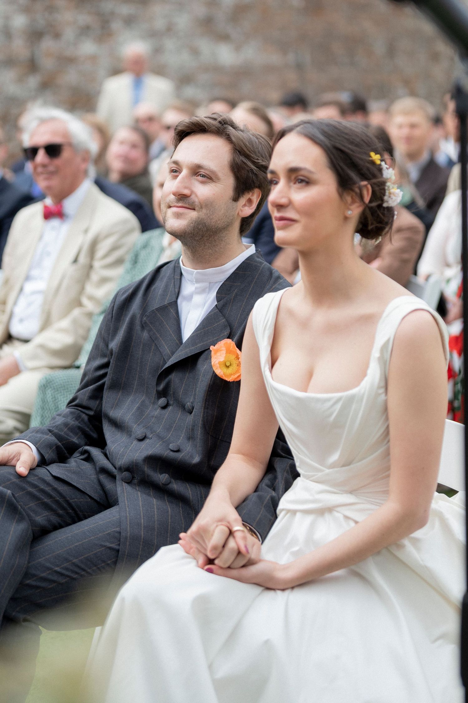 Bride in a white gown and groom in a pinstripe suit with an orange boutonniere sit hand in hand during an outdoor wedding ceremony.