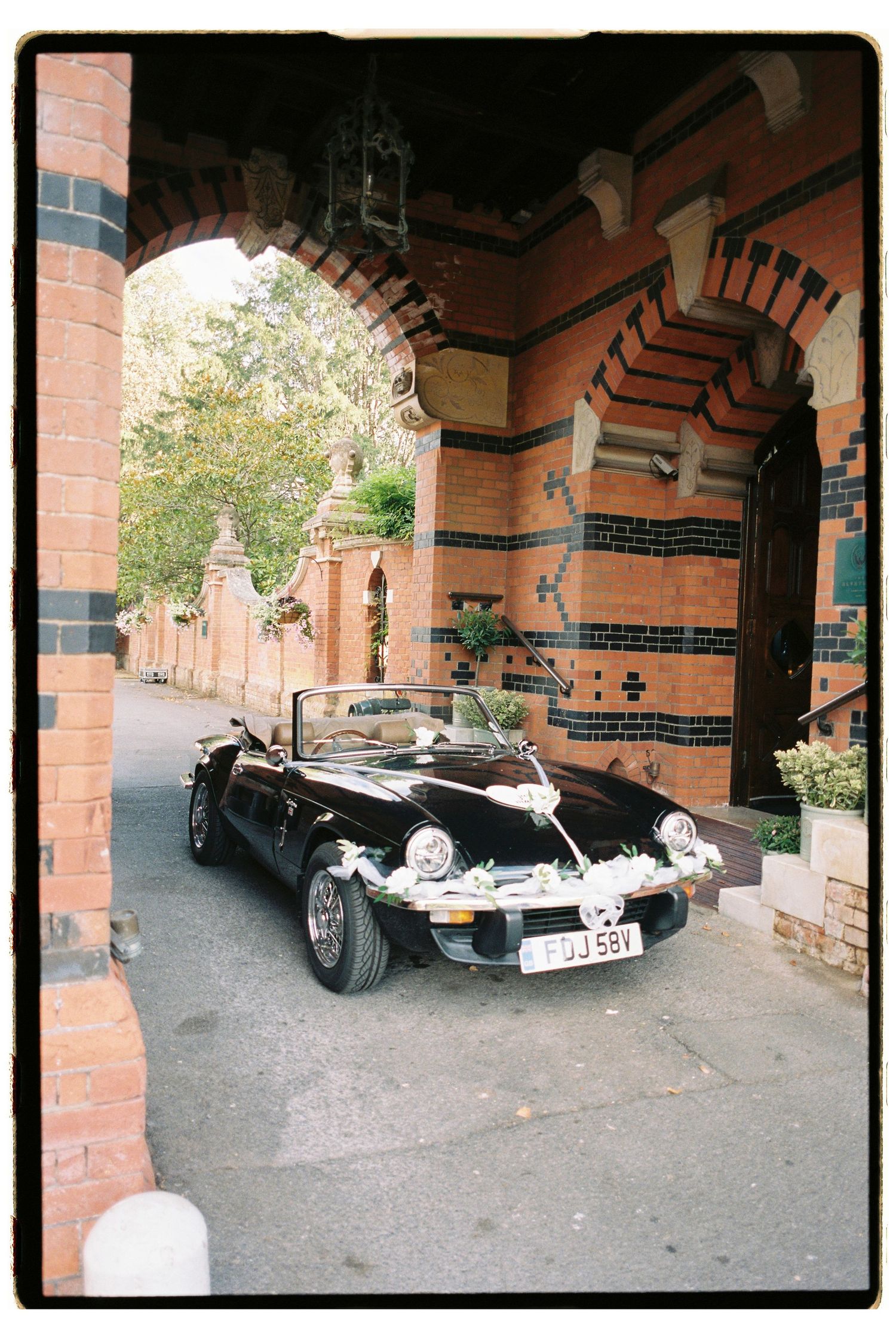 Black vintage convertible wedding car decorated with white ribbons parked outside ornate red brick building entrance.