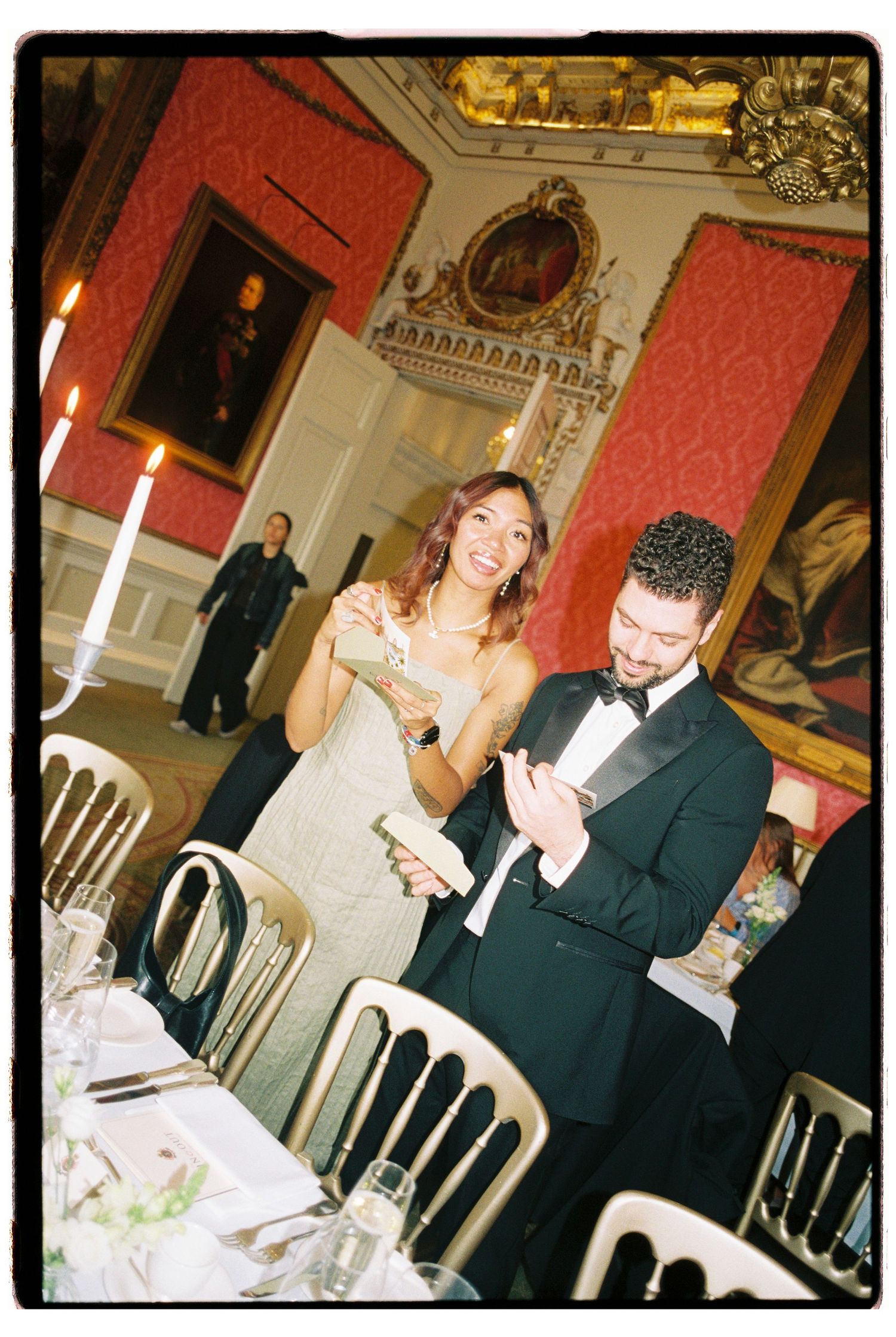 Couple in formal attire laughing together in opulent red-walled banquet hall during elegant wedding reception.
