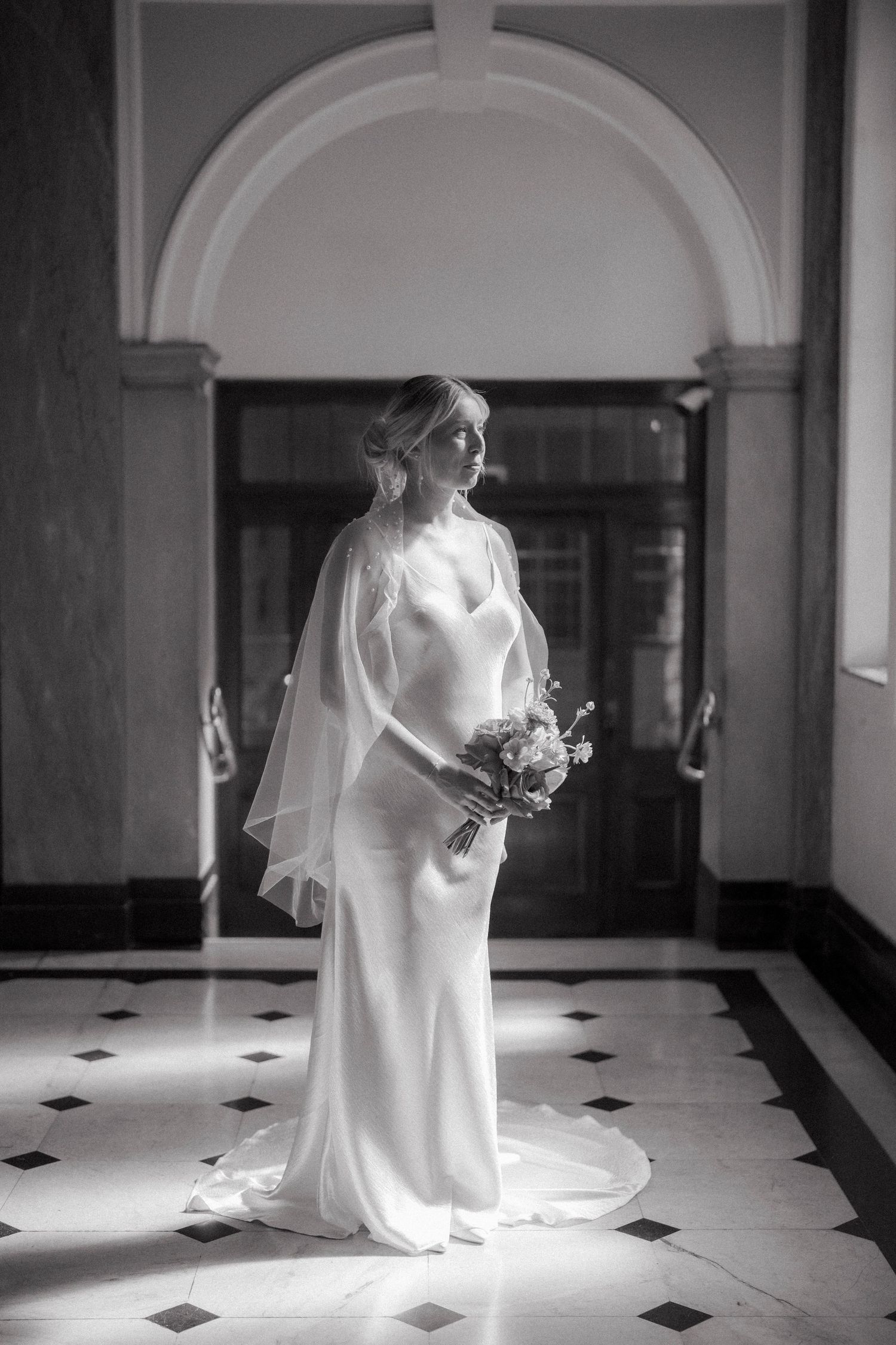 Elegant bride in a sleek white gown and cape veil holds a bouquet in a grand hall with arched doorway.