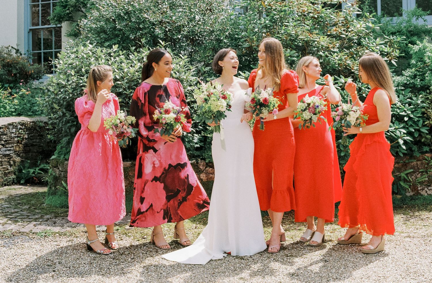 Bridesmaids in coral dresses pose with bride in white on garden grounds.