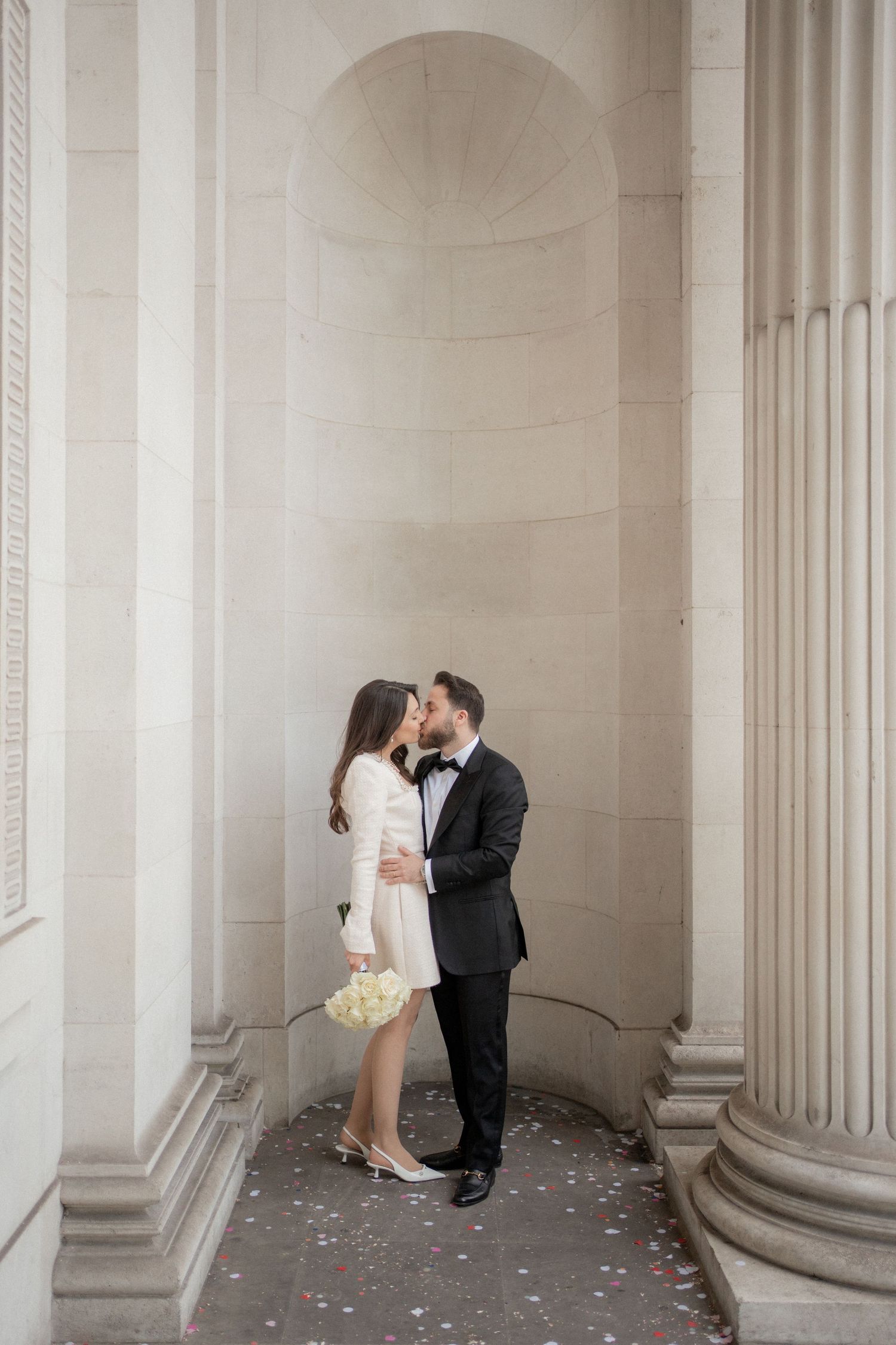 A romantic wedding couple shares intimate moments between elegant stone columns in a classic architectural setting.
