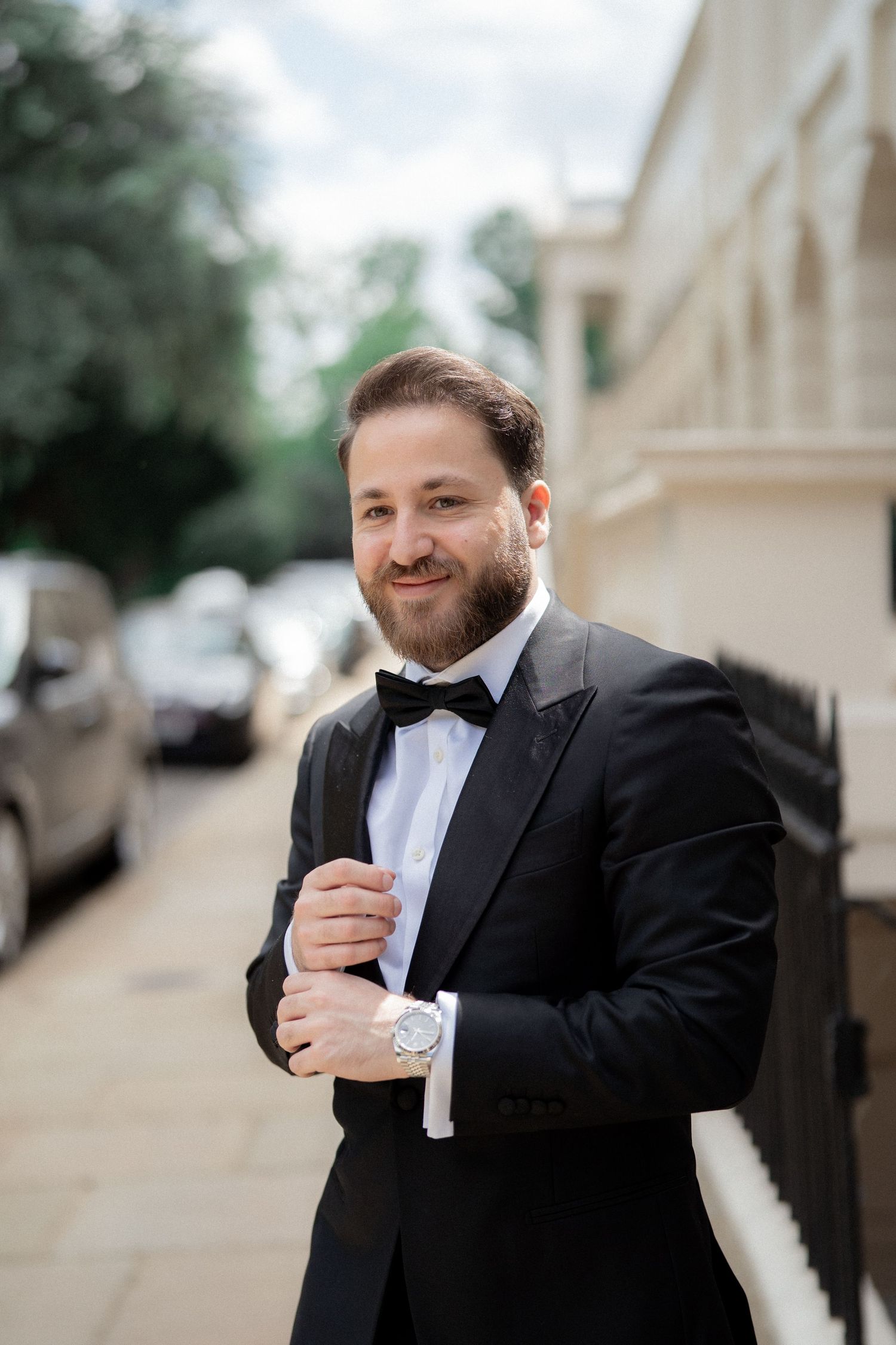 A person in a black tuxedo and bowtie adjusts their sleeve while posing outdoors.