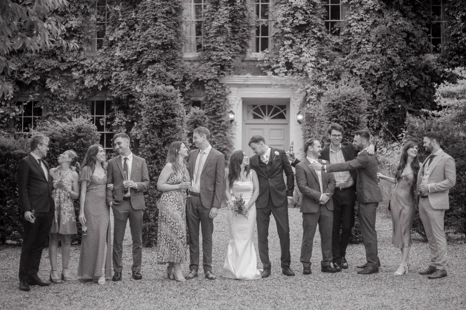Black and white wedding photo of bride, groom, and guests posing outside an ivy-covered manor house.