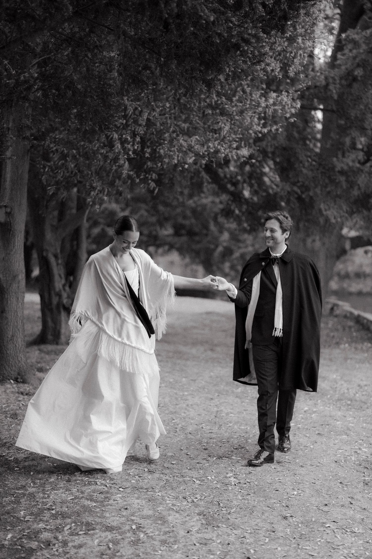 Black and white photo of a bride in a flowing white fringed gown holding hands with a smiling groom in a dark cape on a tree-lined path.