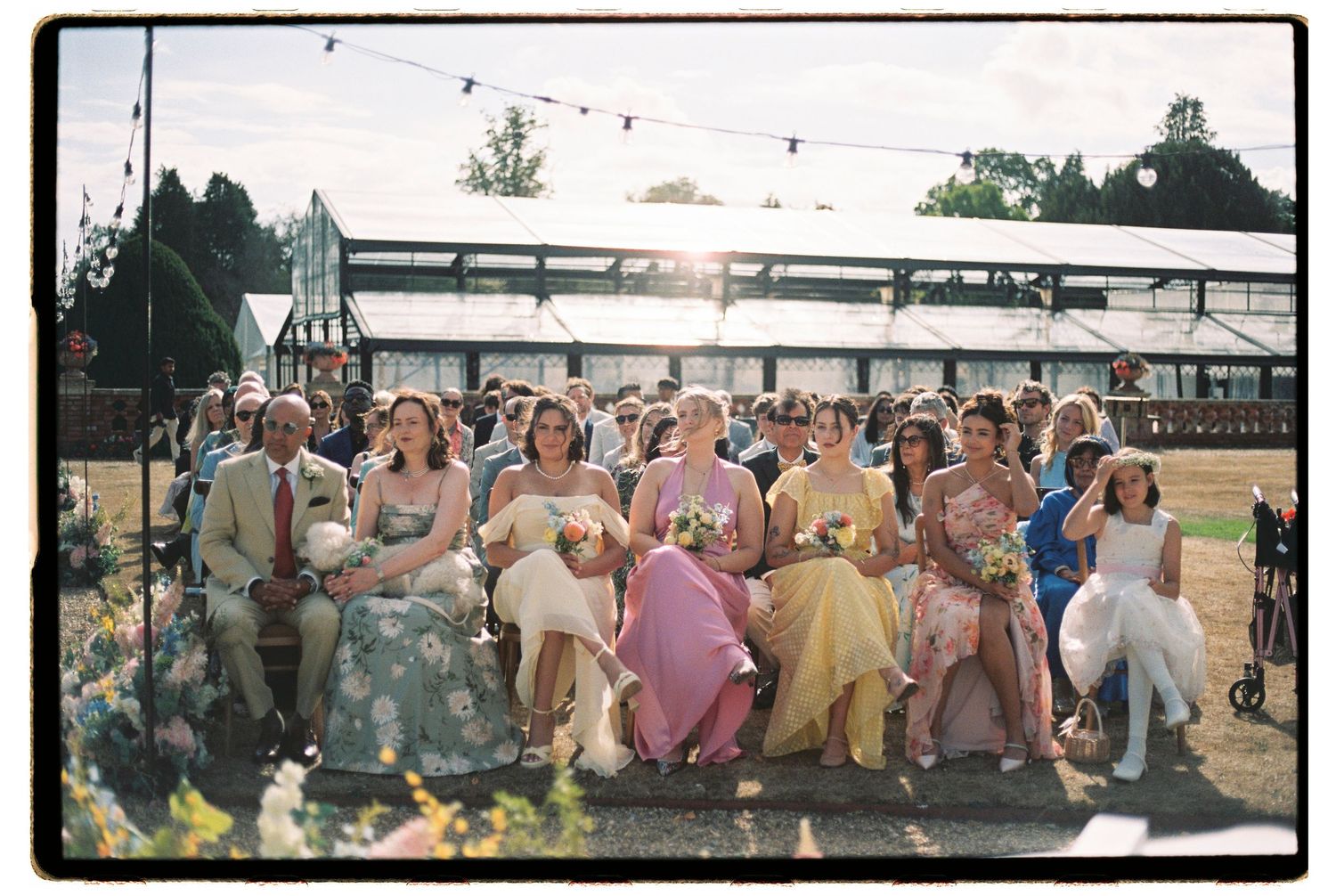 Colorful wedding guests in floral and pastel dresses seated outdoors at a sunny garden ceremony.
