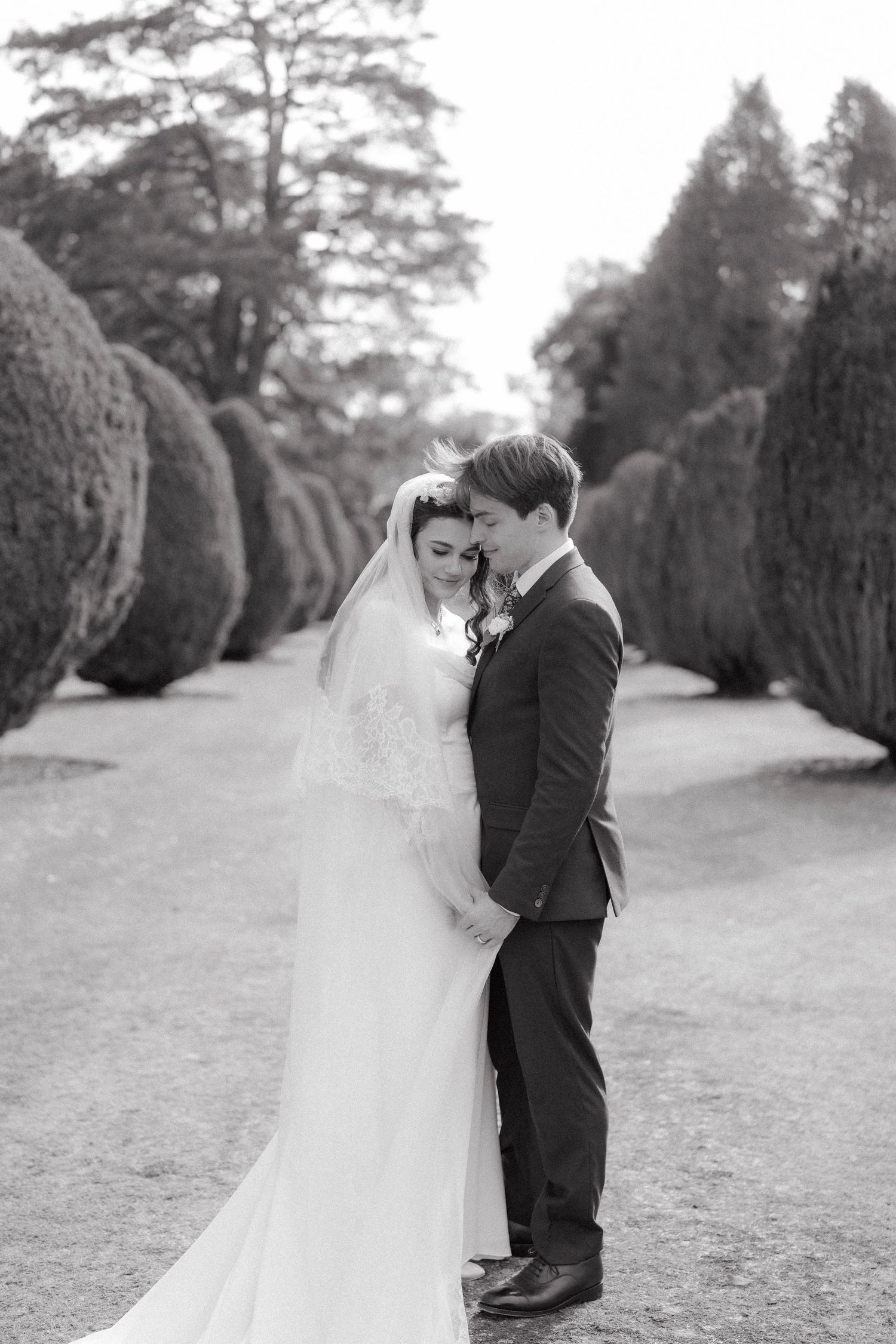 Black and white photo of bride and groom embracing on a tree-lined path at their elegant wedding.