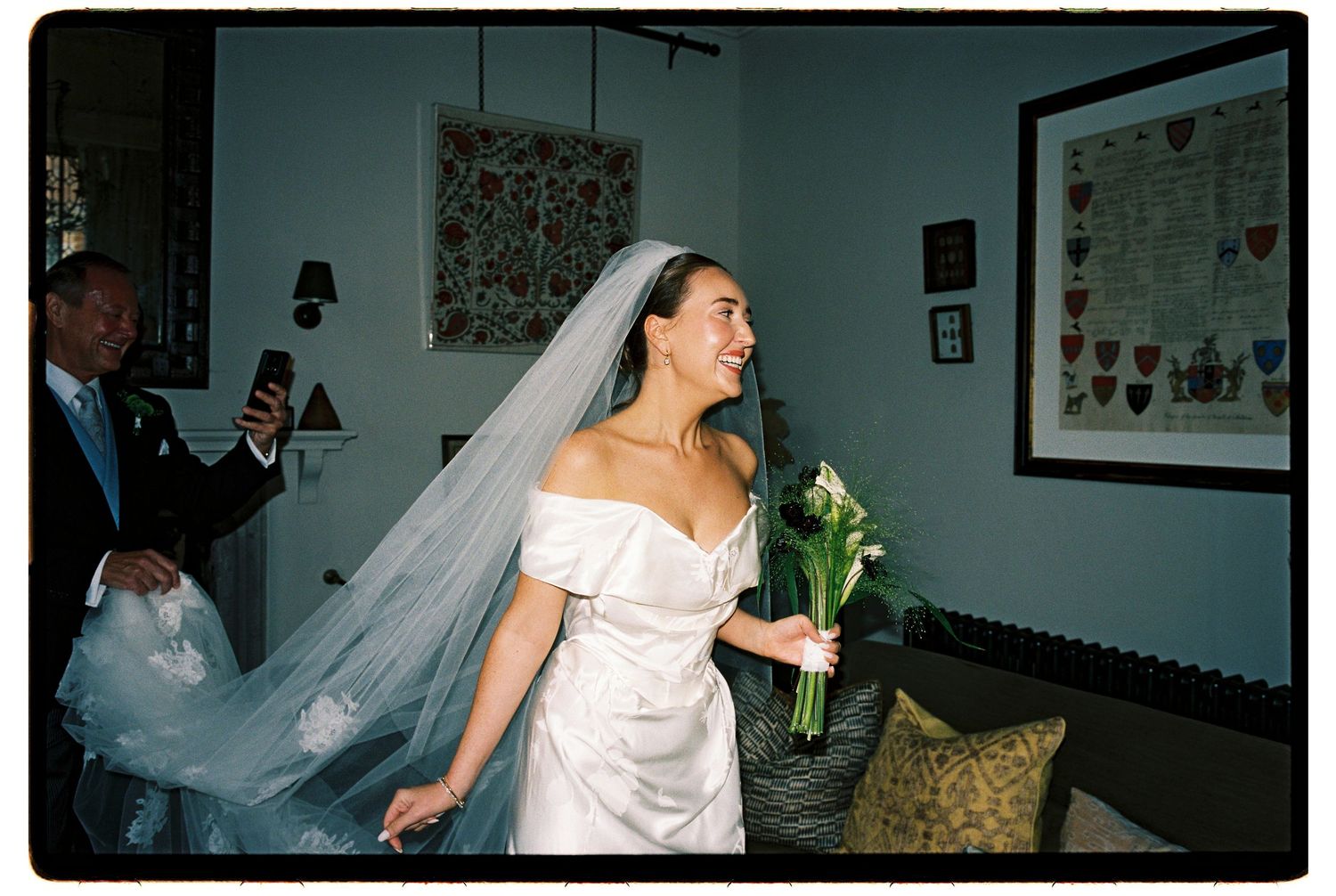 Smiling bride in off-shoulder white gown and veil holds calla lily bouquet indoors while man photographs her.