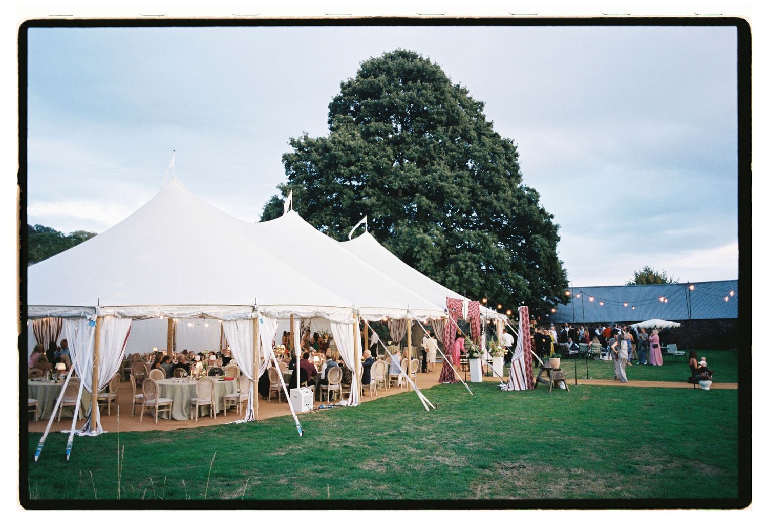 Elegant white pole marquee wedding reception tent glowing at dusk on lush green countryside lawn.