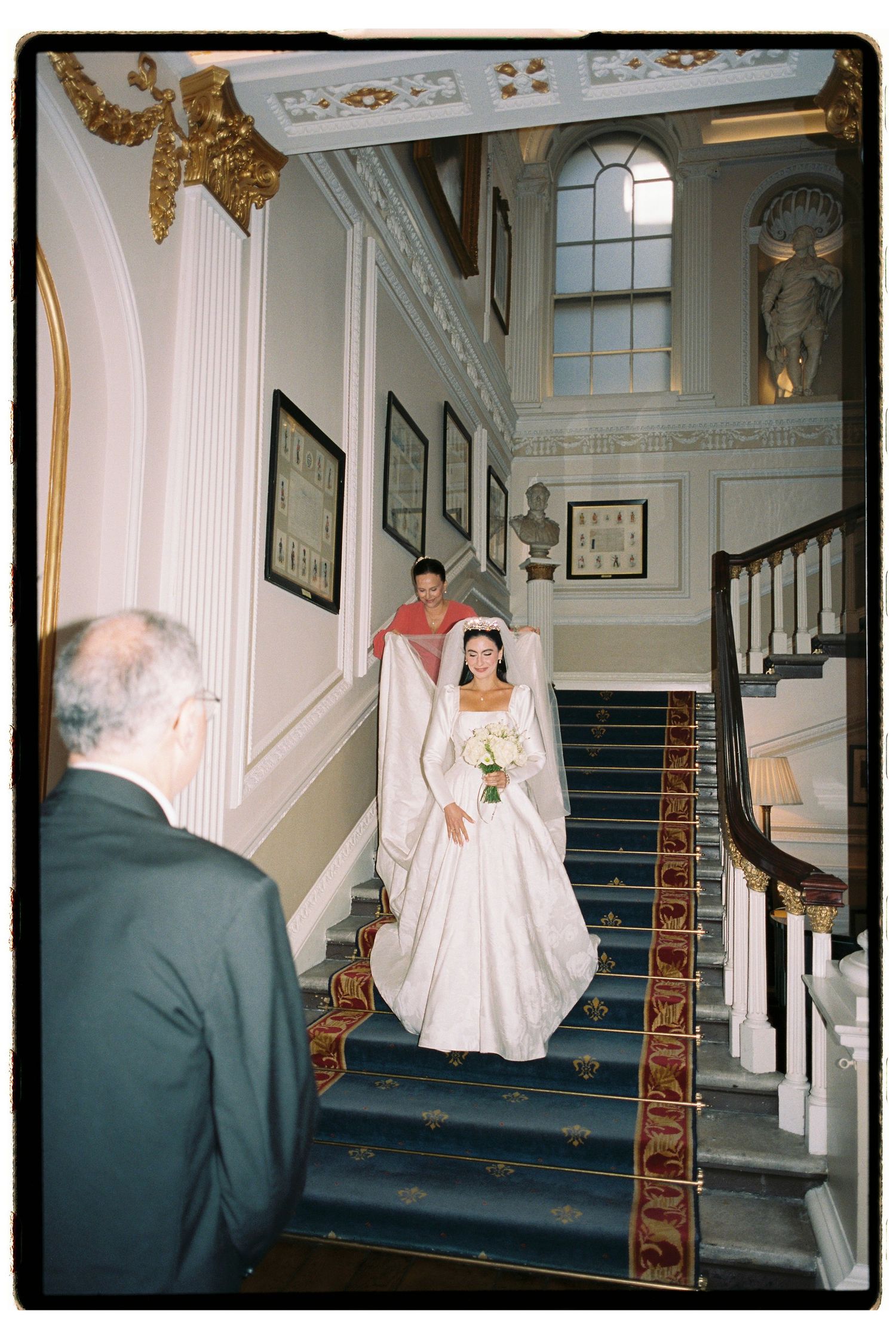 Bride in white wedding gown descends grand staircase with escort at elegant venue.