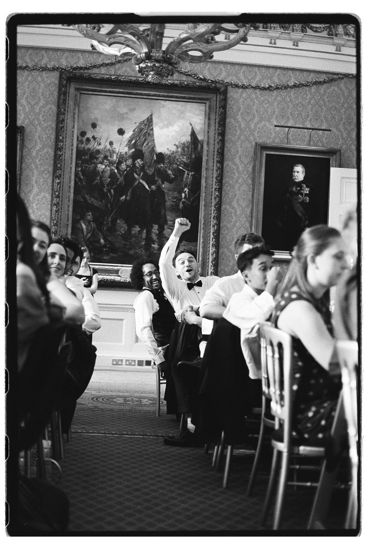 Wedding guests celebrate at reception in ornate room with historical painting, one guest raises fist in excitement.