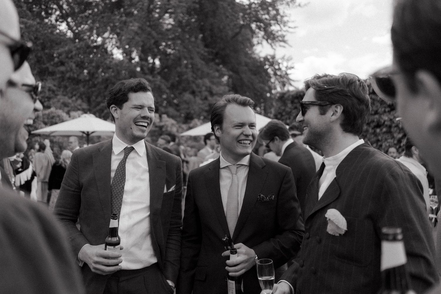 Three smiling men in suits hold drinks and chat at an outdoor wedding reception under trees in black and white.
