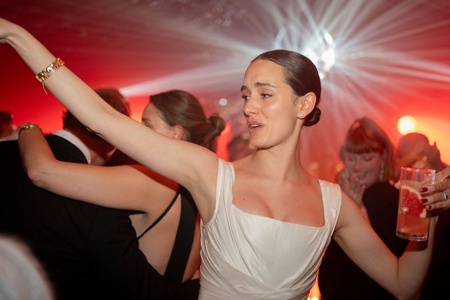 Bride in a white dress dances with arms raised on a crowded dance floor beneath glowing red lights at a lively wedding reception.