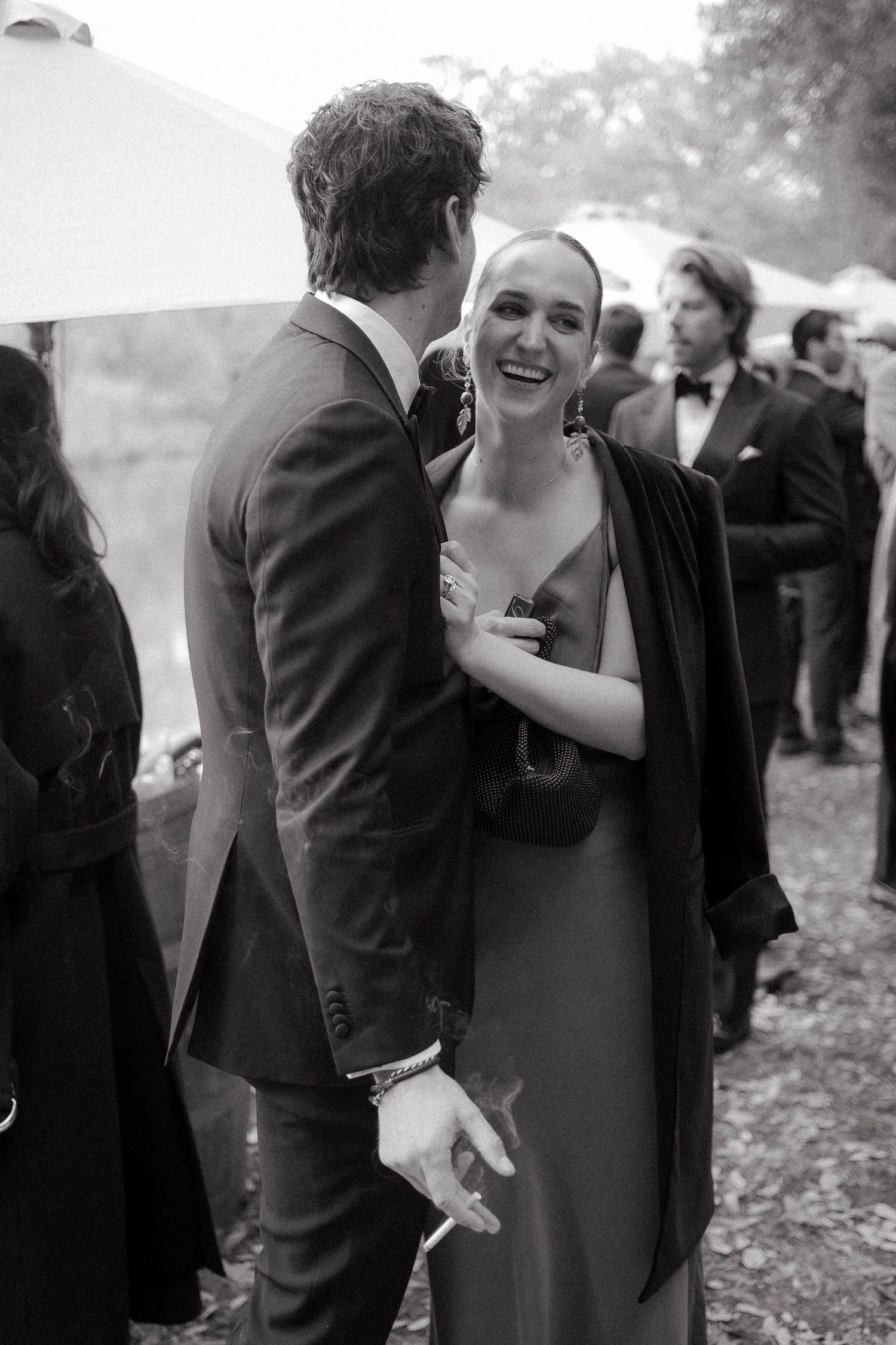 Black and white candid of a laughing bride in a satin slip dress and dark jacket chatting with her groom at an outdoor wedding reception.