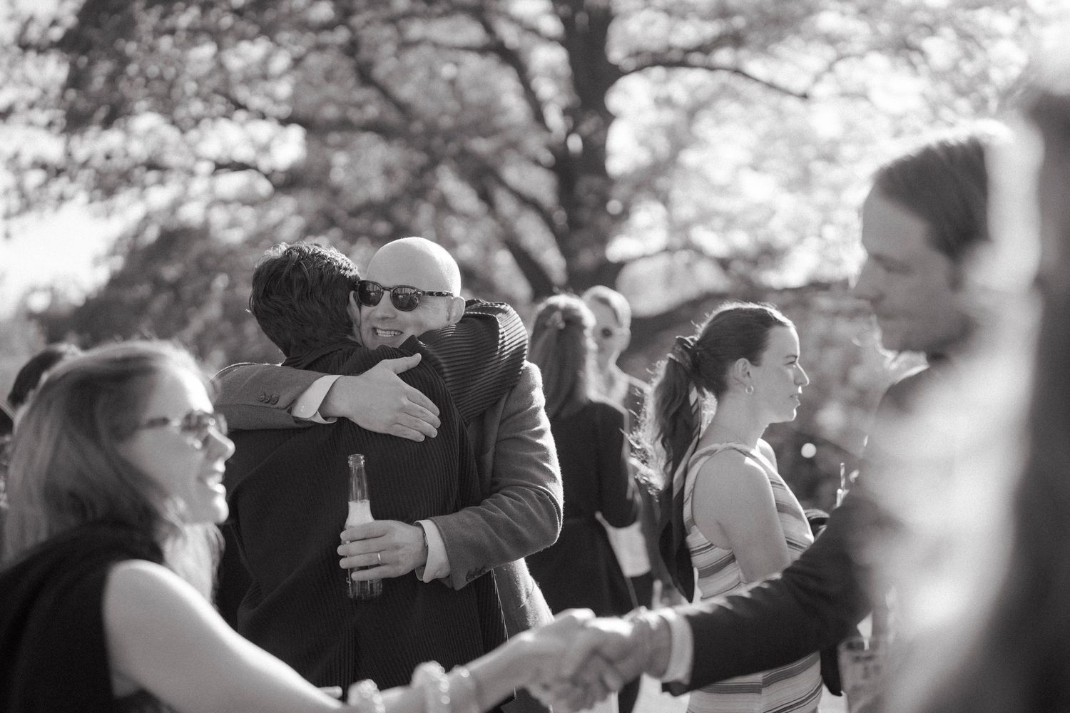 Wedding guests share a warm embrace in a black and white photo, with friends smiling and shaking hands under leafy trees.