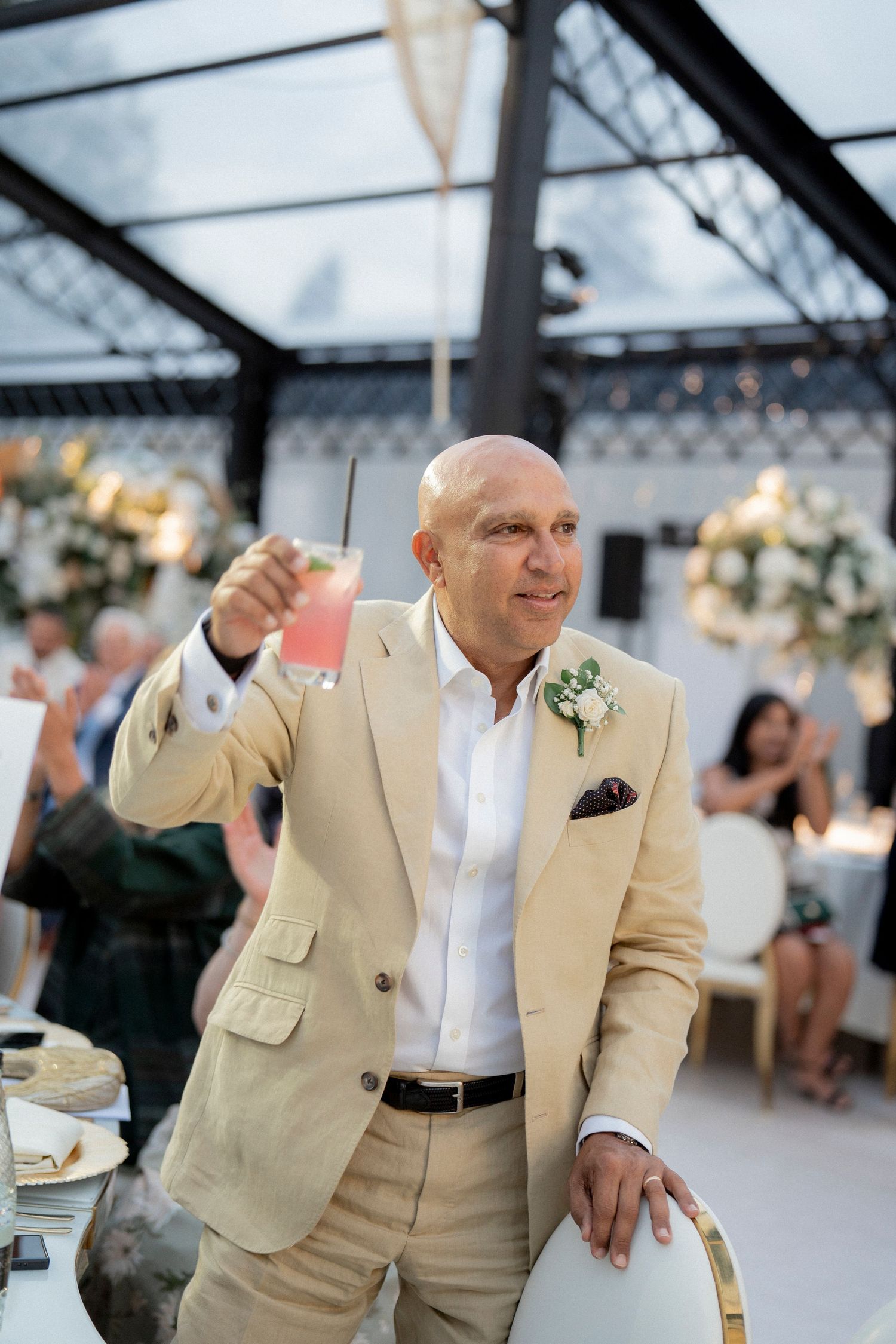 Bald man in beige suit raises a pink cocktail toast at an elegant wedding reception.
