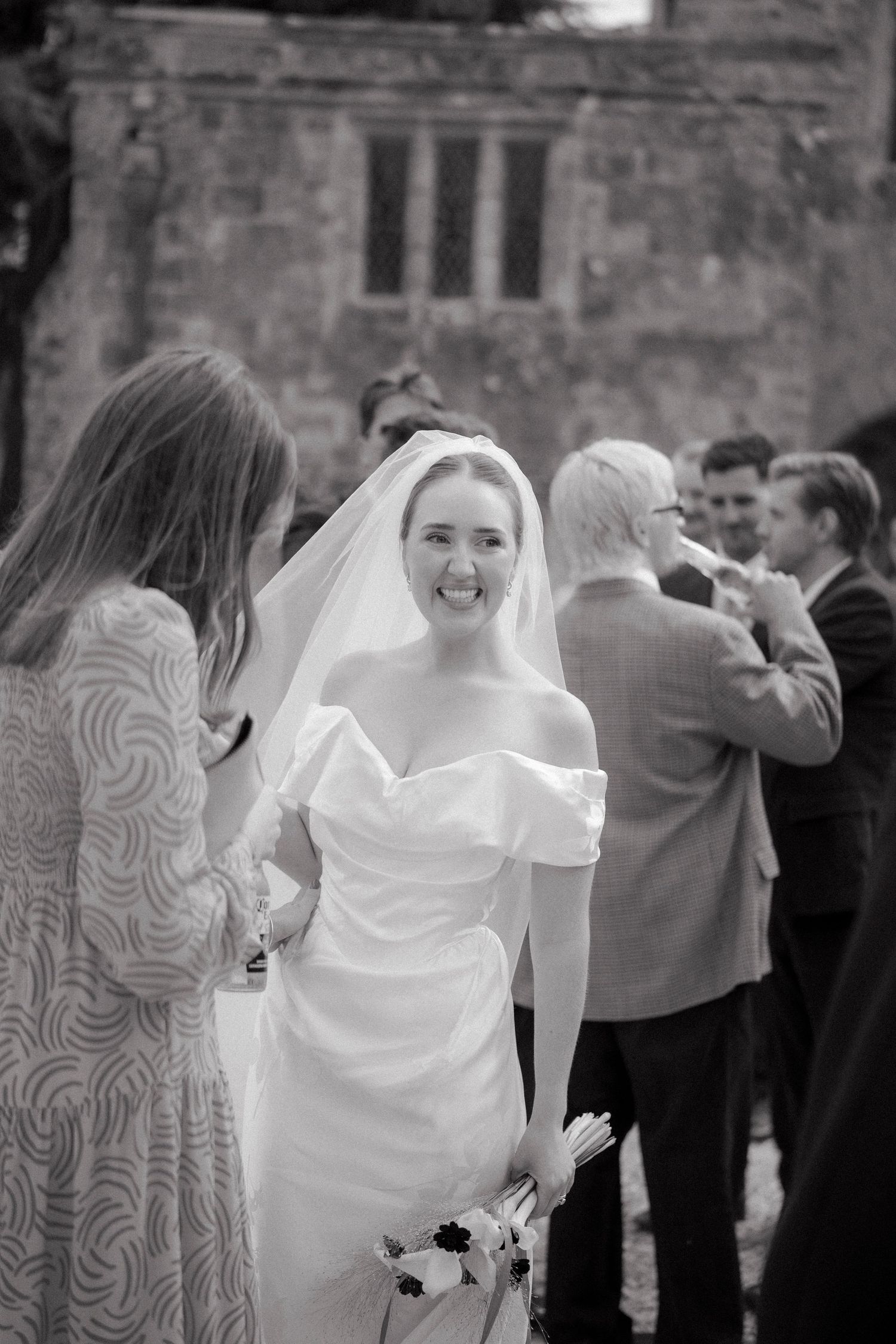 Smiling bride in white off-shoulder gown and veil mingles with guests outside a historic stone church.