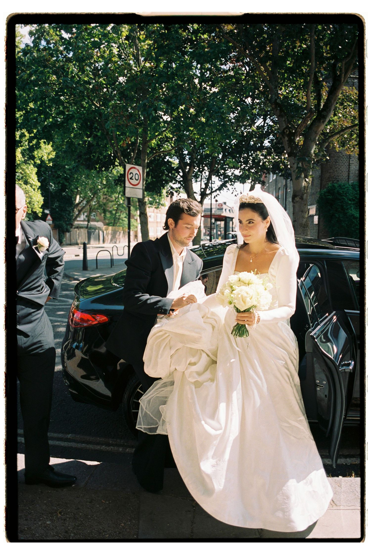 Bride in white gown holding bouquet exits black car assisted by groom in suit on a sunny street.