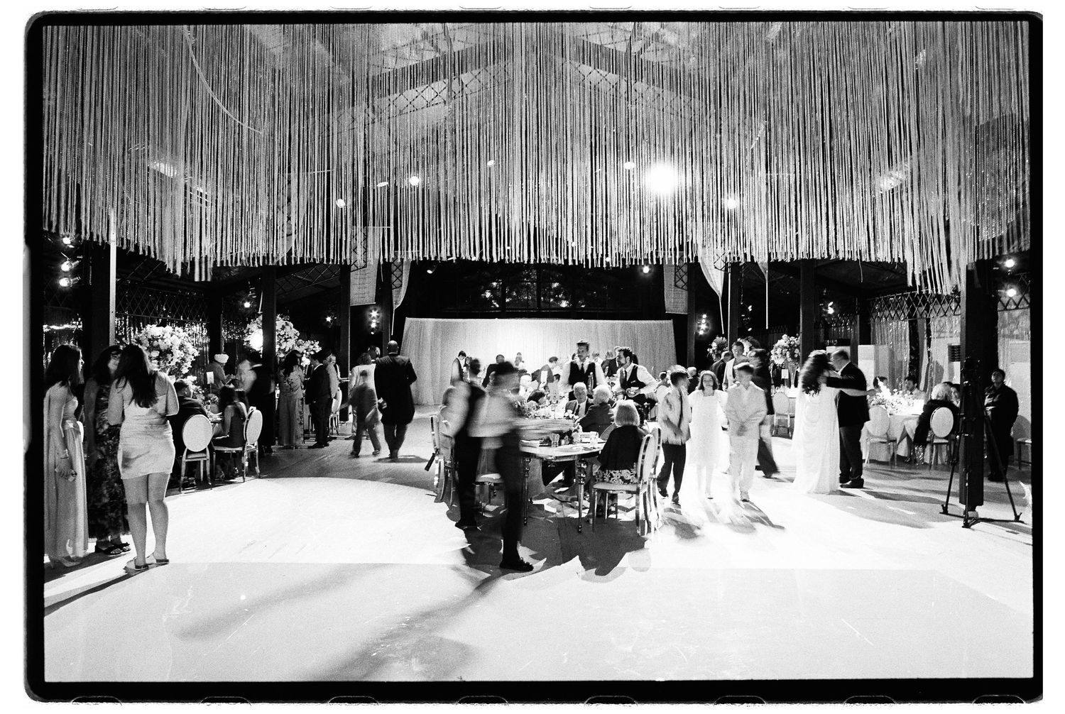 Black and white photo of elegant wedding reception with hanging streamers, guests mingling on dance floor under festive lighting.