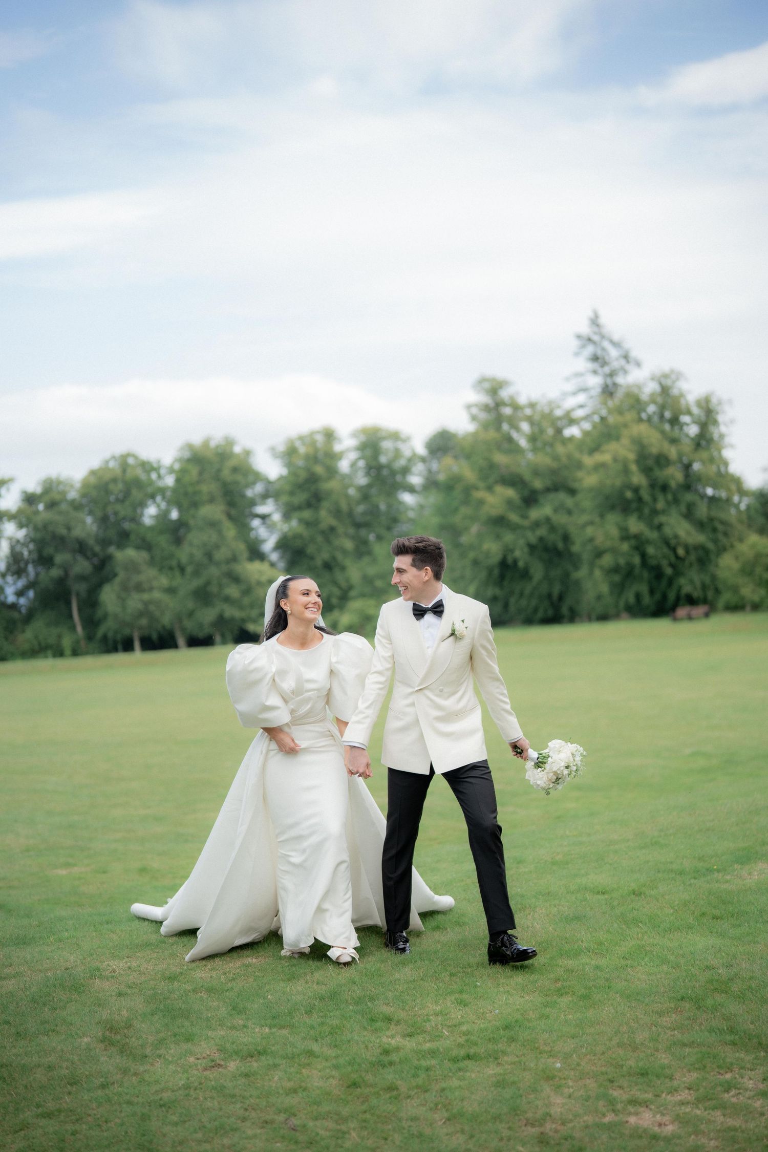 Wedding couple in formal attire playfully posing on a green lawn under blue skies.