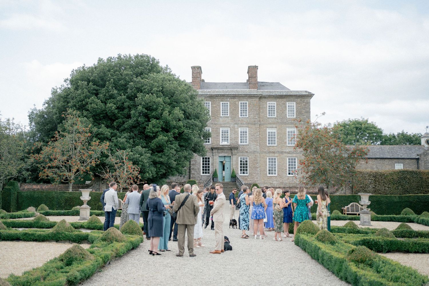 Guests gather in formal garden of elegant Georgian manor house for outdoor wedding reception.