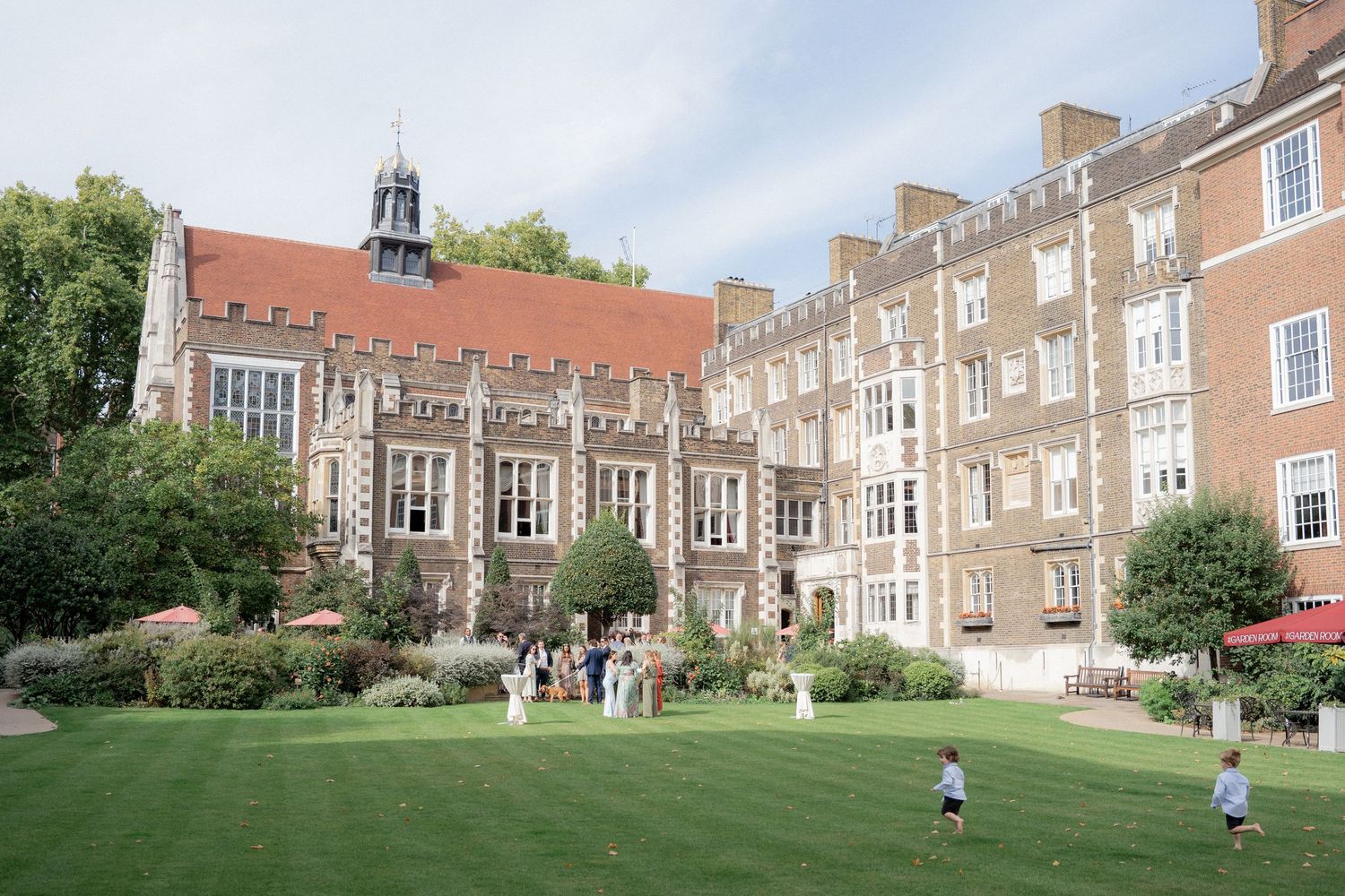 Historic brick university courtyard with lush green lawn, Gothic architecture, and visitors enjoying a sunny day.