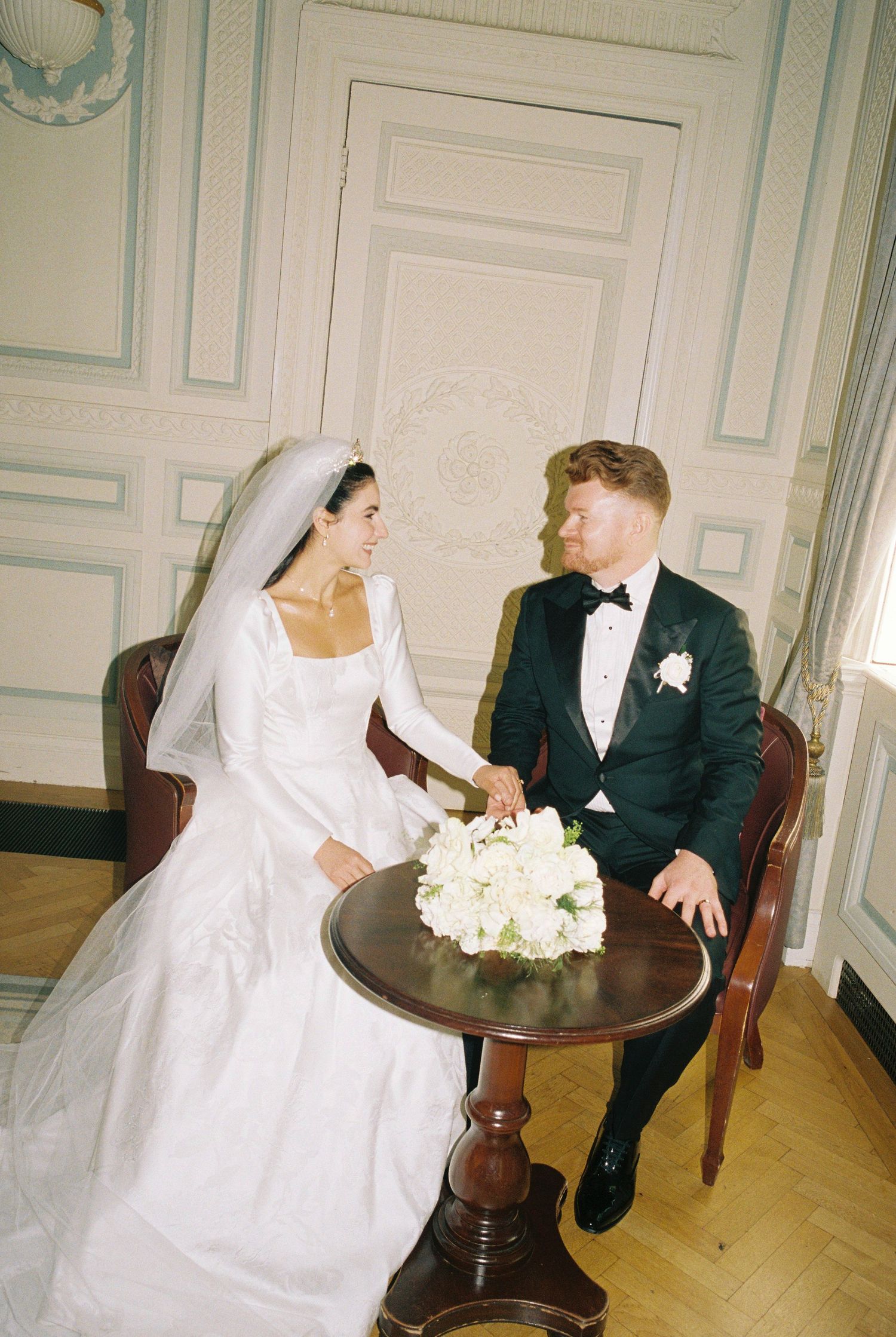 Bride in a white long-sleeved gown and veil sits smiling at a groom in a black tuxedo beside a small table with white flowers.
