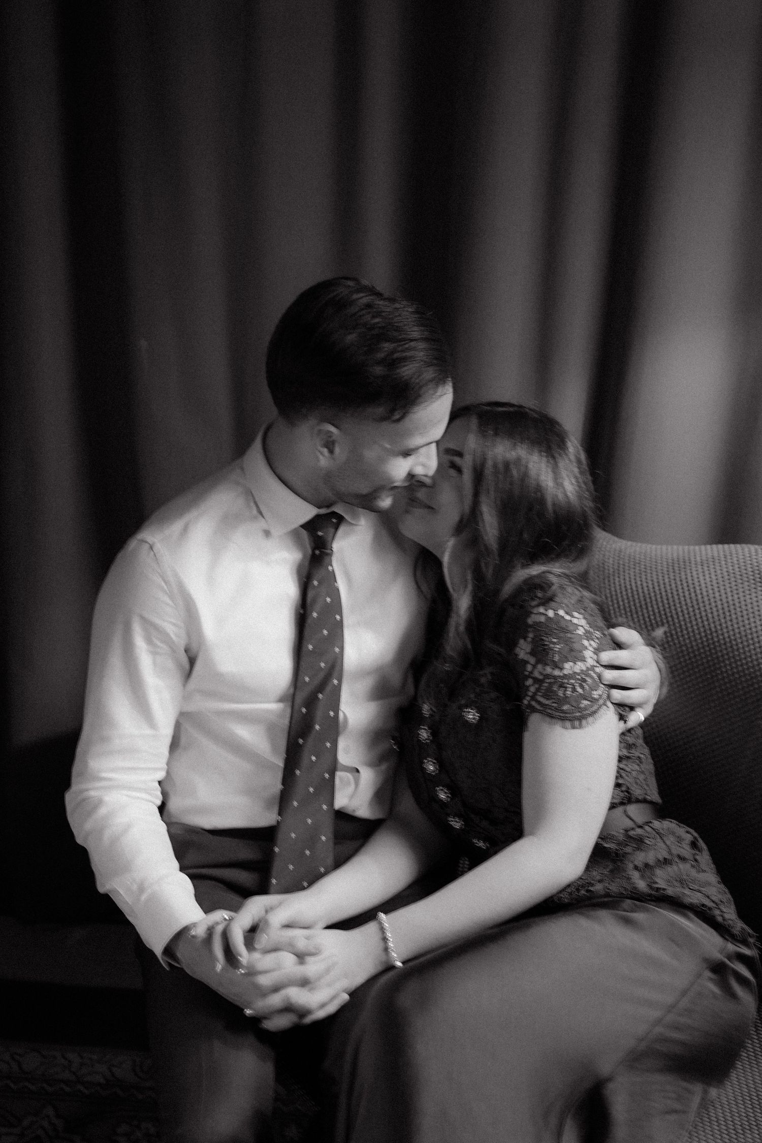 Black and white photo of a couple sharing a tender moment, sitting close together on a chair, smiling at each other.