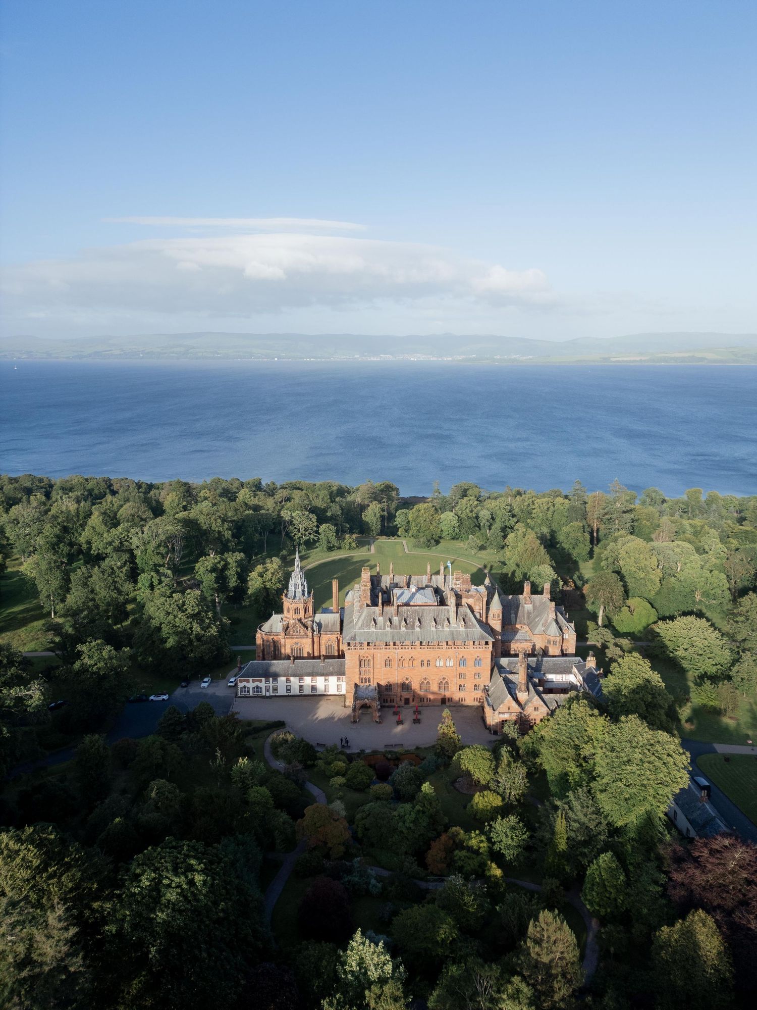 Aerial view of a grand red brick castle surrounded by lush green trees with a vast blue sea in the background.