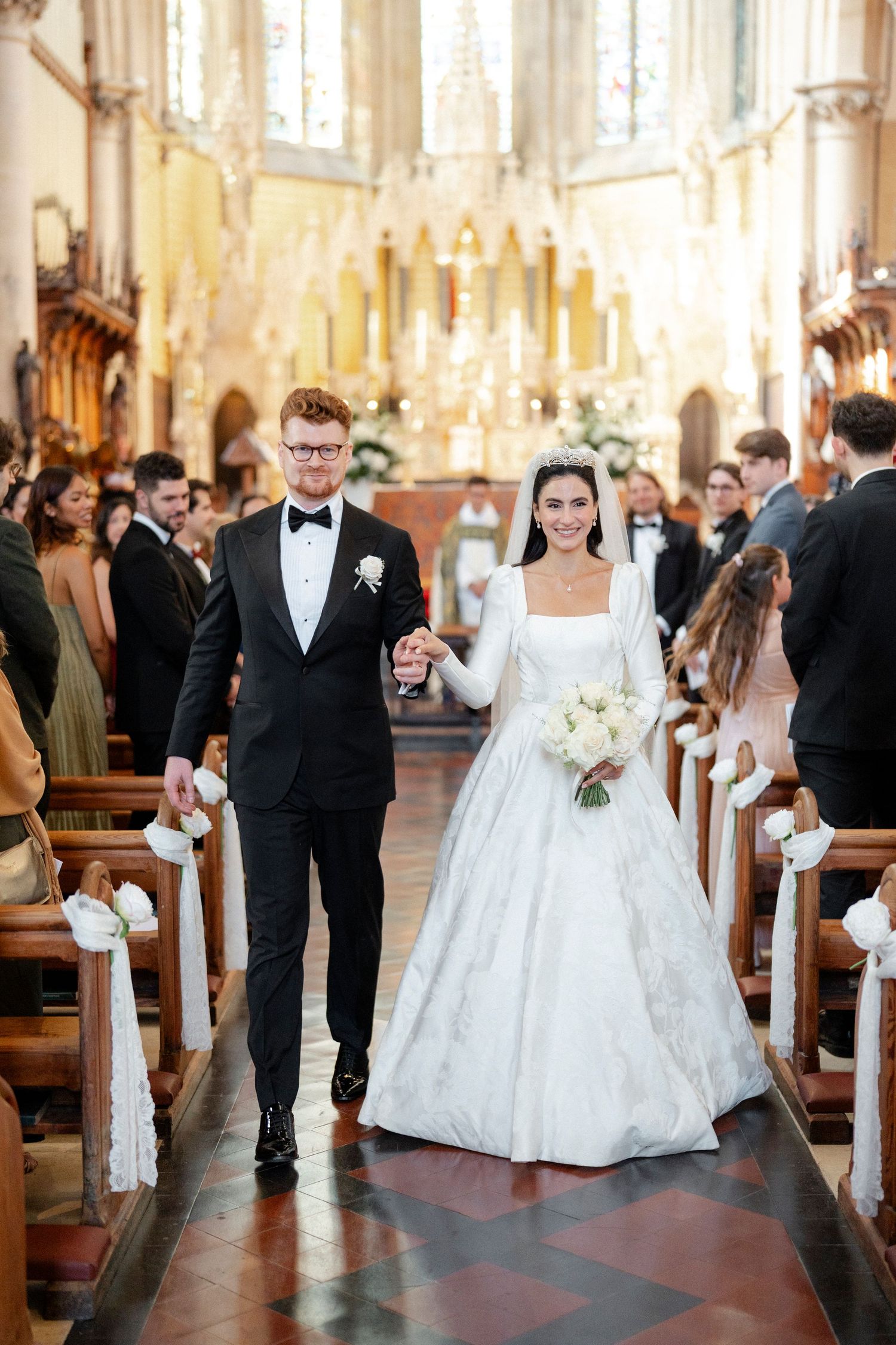 Bride and groom walk down church aisle, she holds white bouquet in elegant long-sleeve gown.