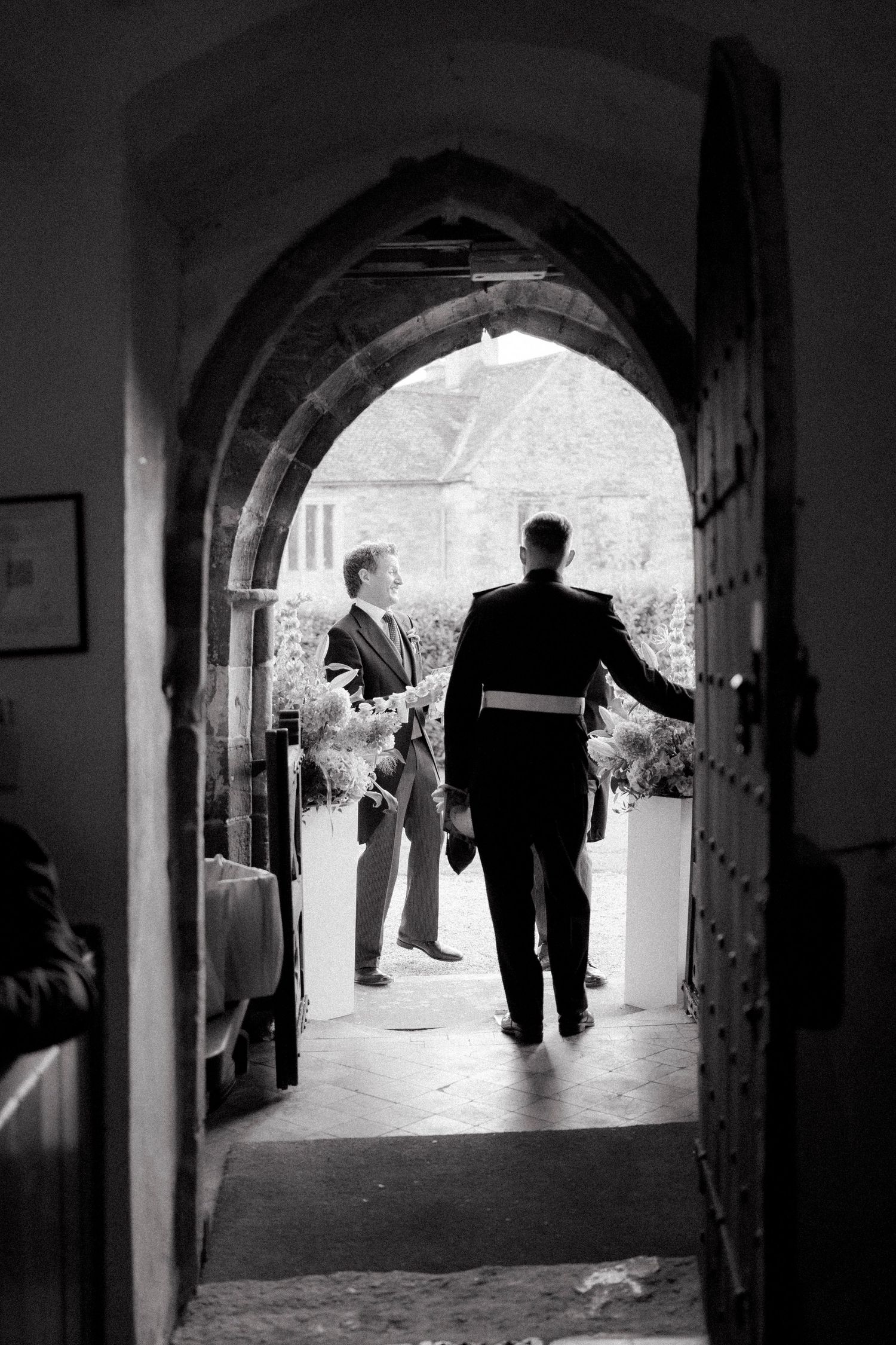 Two men greet each other through a Gothic stone church archway during a wedding ceremony.