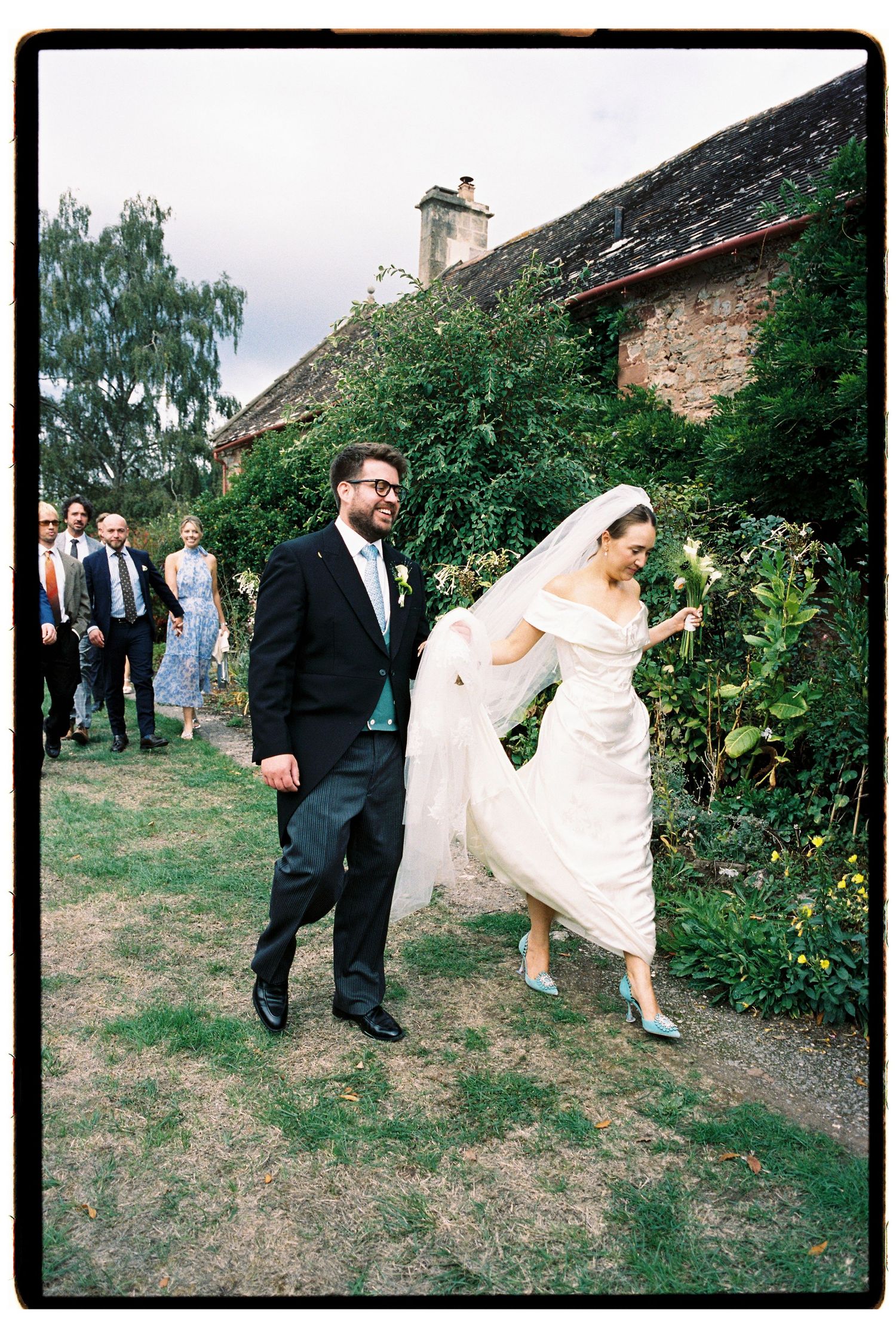 Bride in white gown and veil with groom in black suit walking outdoors at a garden wedding venue.