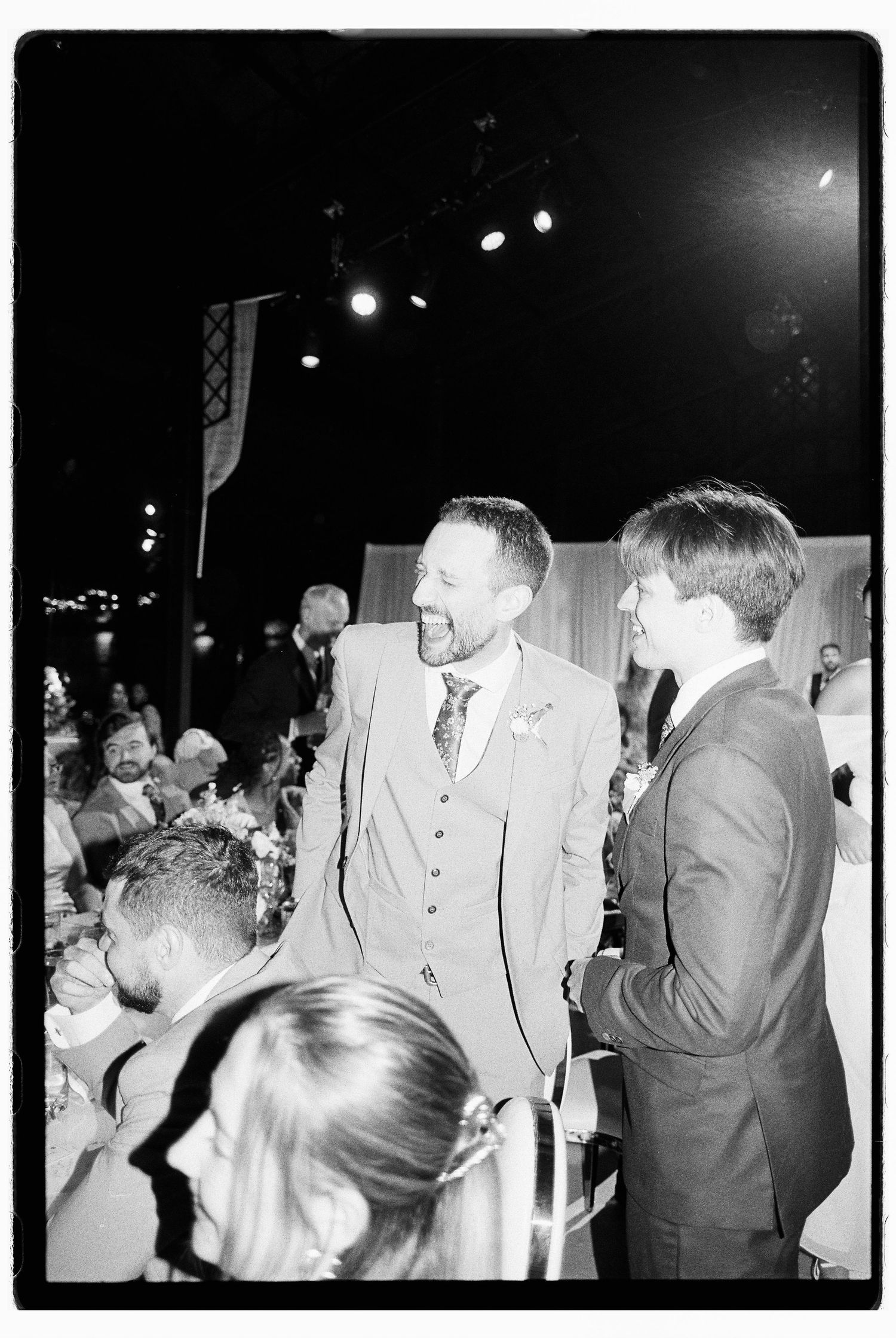 Groom in three-piece suit laughs with guest during wedding reception in black and white candid photo.