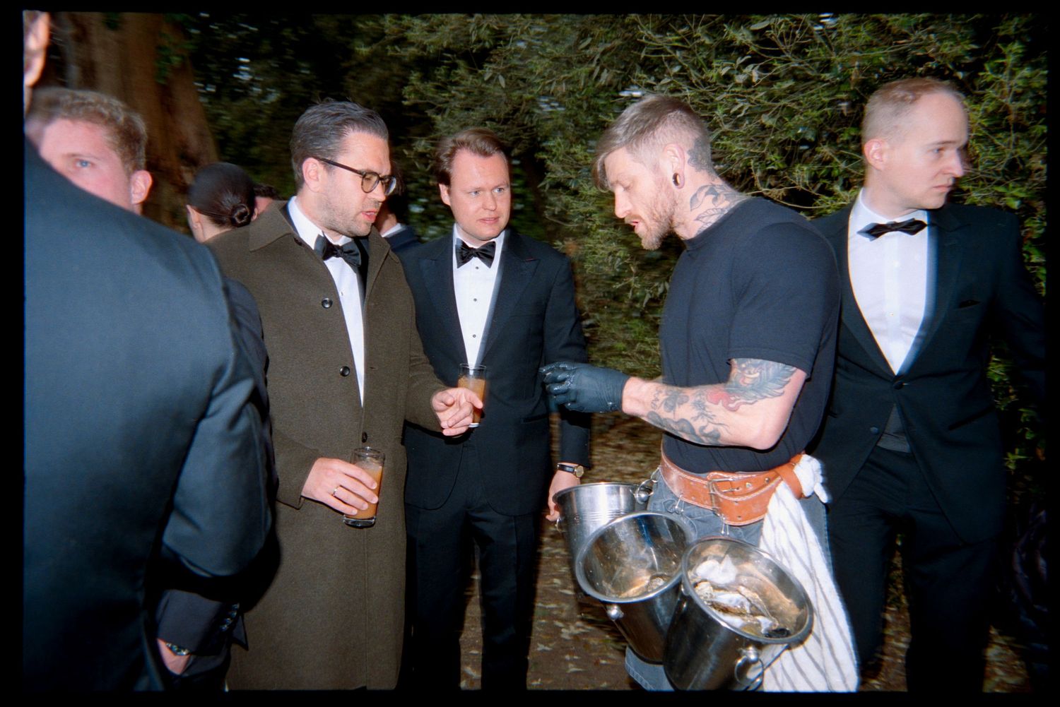 Guests in black tie watch a tattooed man in an apron stir a metal pot at an outdoor wedding reception.