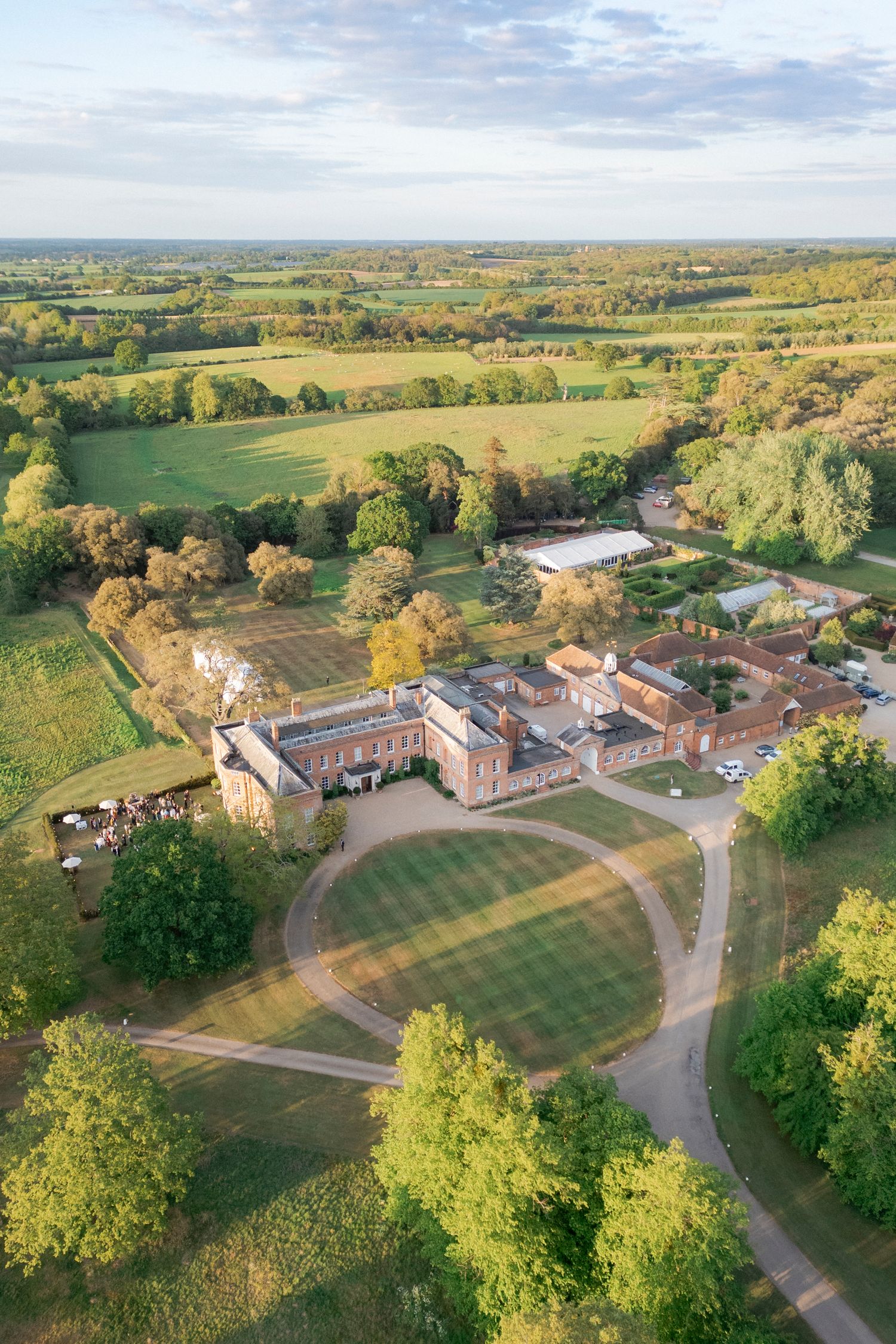 Aerial view of a red brick country manor with a circular driveway, manicured lawn and surrounding countryside fields and trees.