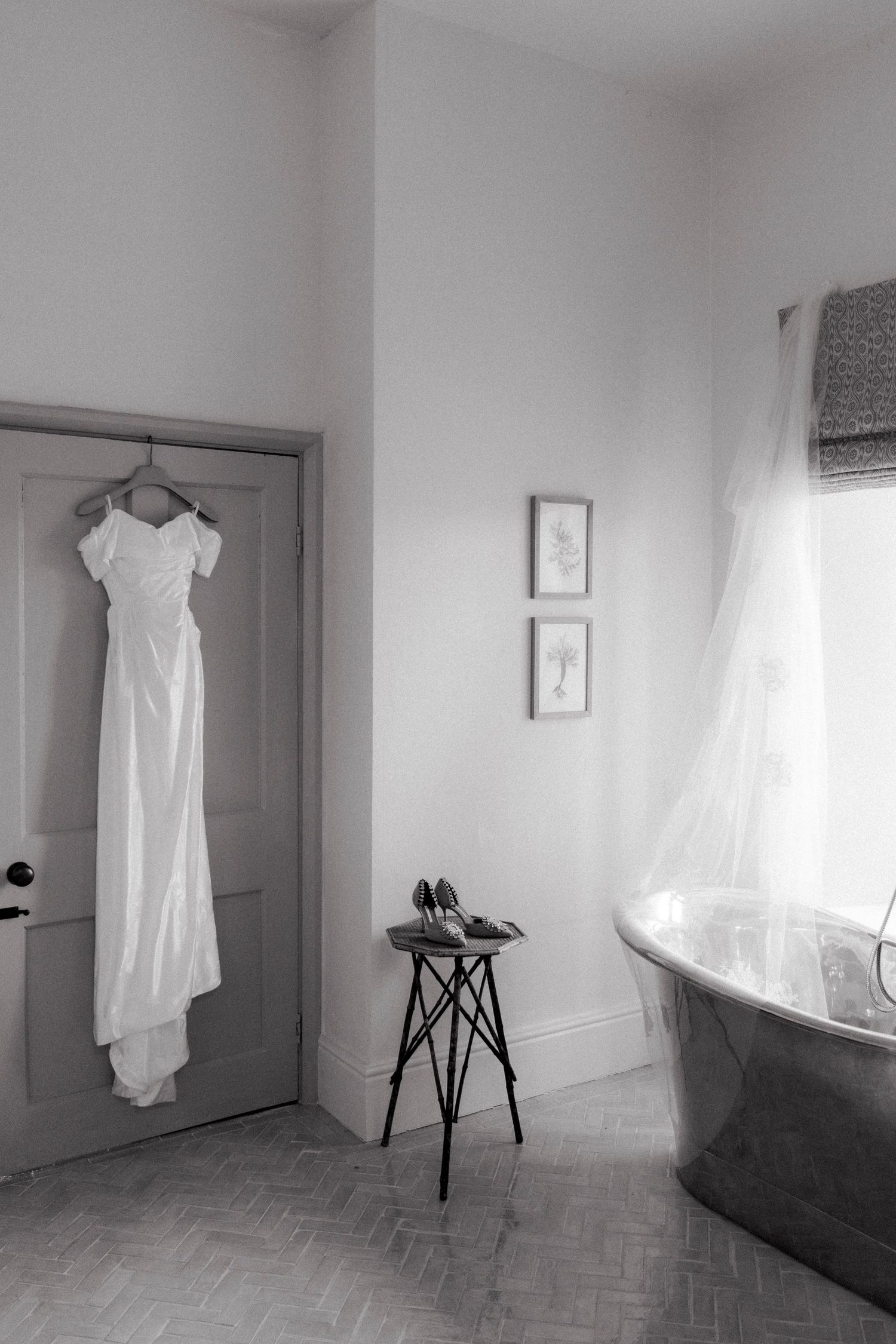White off-the-shoulder wedding dress hanging on a door beside a small stool with bridal shoes and a freestanding tub in a bright bathroom.